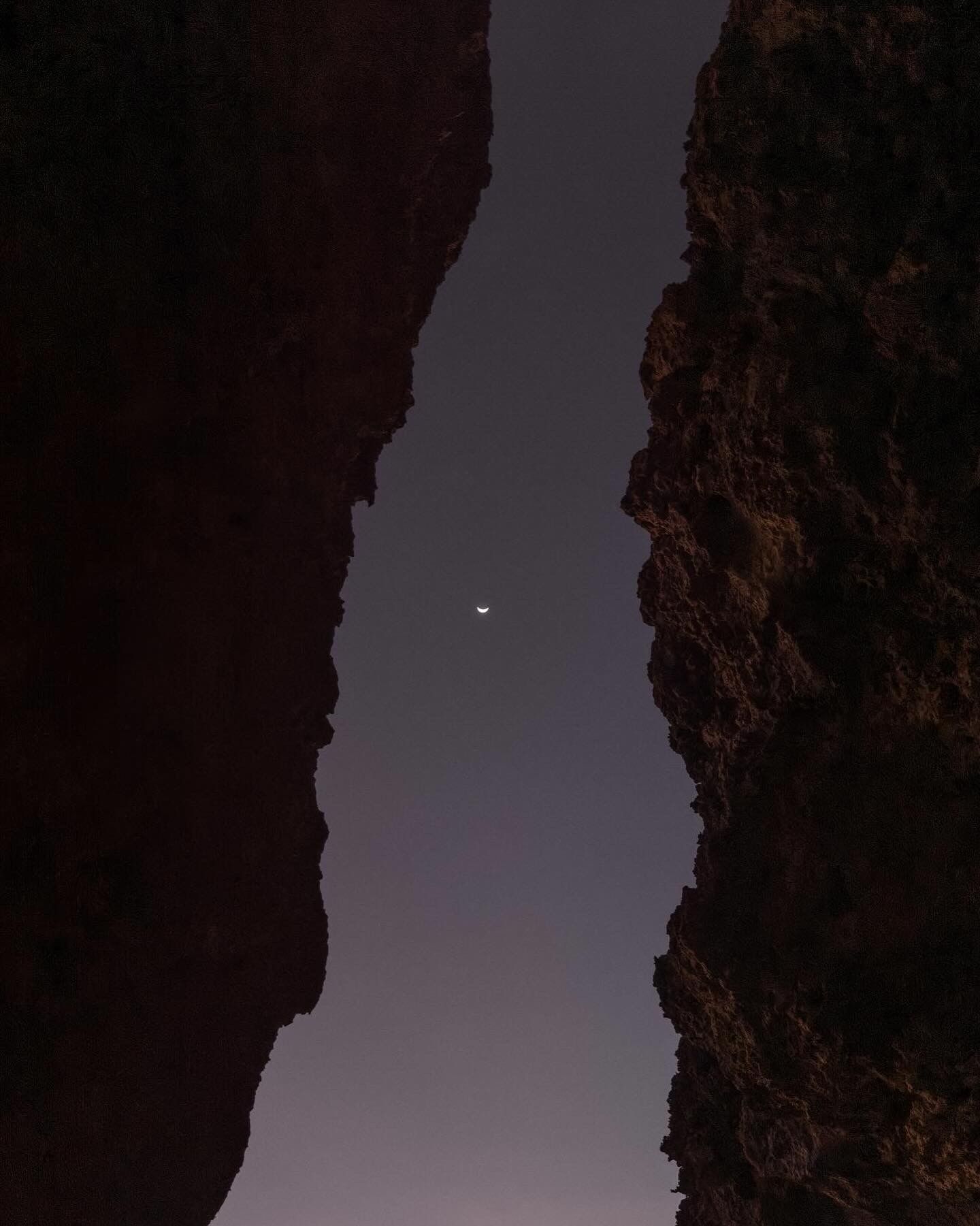 Narrow opening between dark rock cliffs revealing soft twilight sky and distant moon. Bali, Indonesia