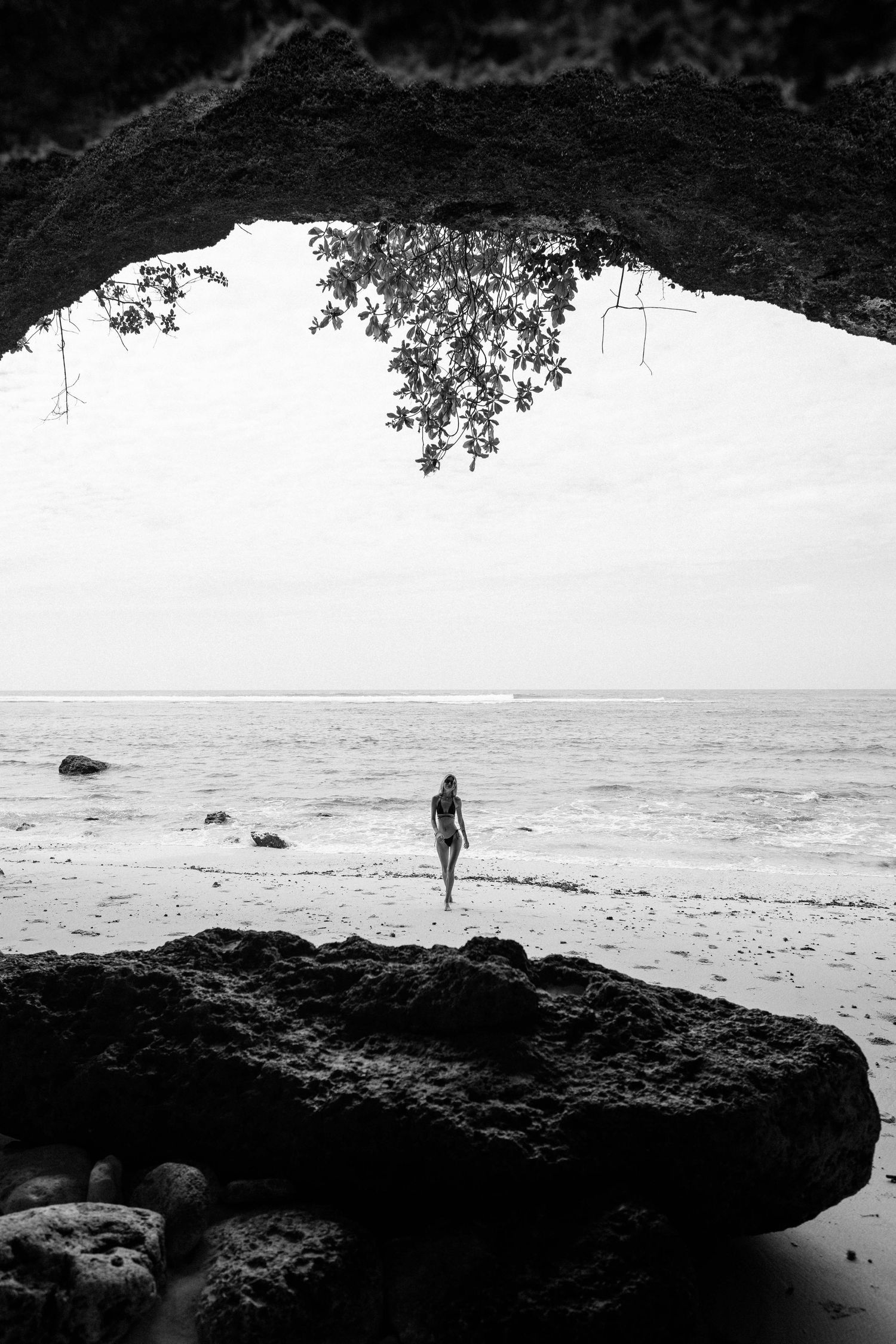 Black and white view from rocky cave opening toward quiet beach and ocean.