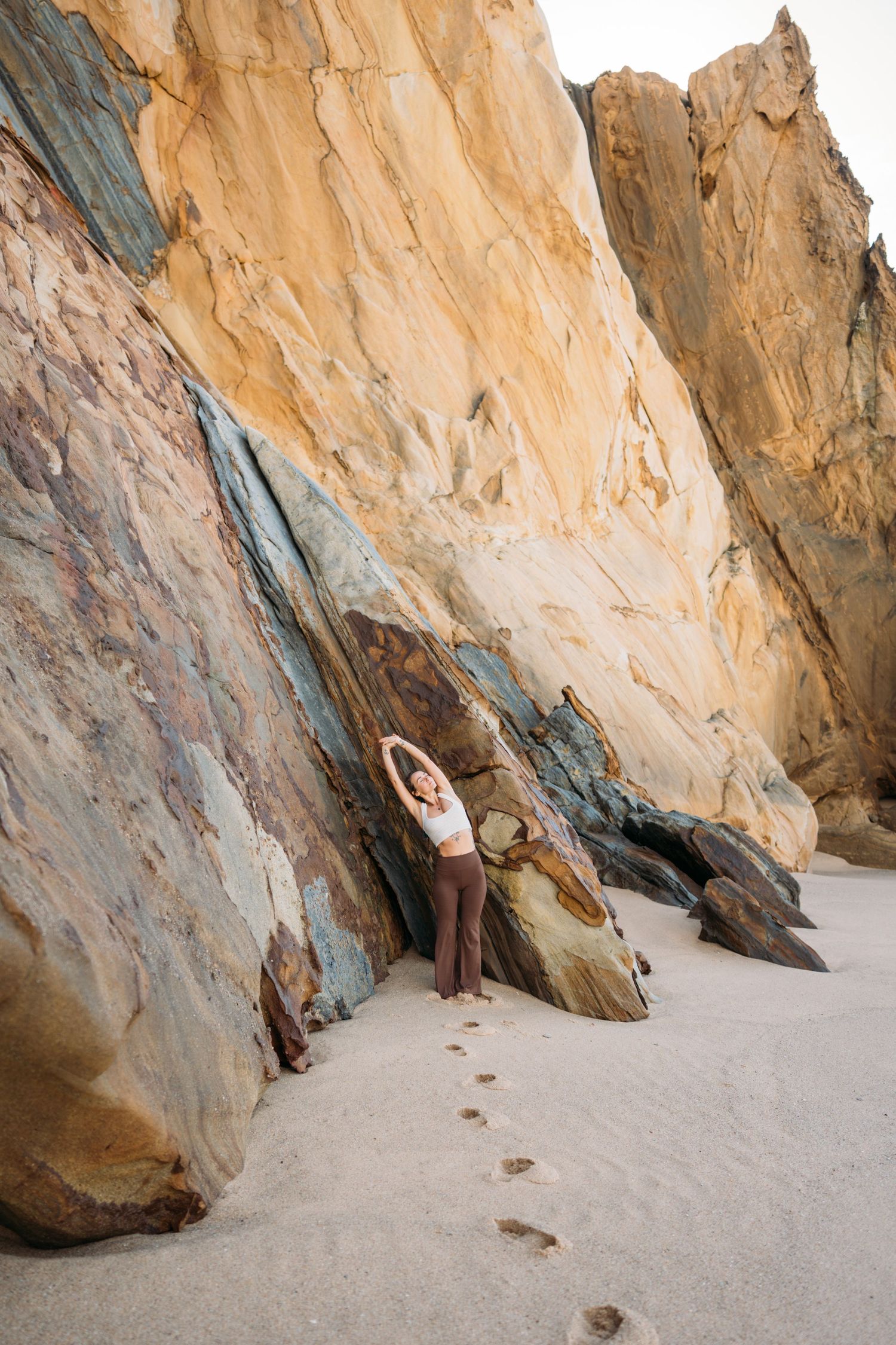 Woman standing beside tall sandstone rock wall on quiet sandy beach.