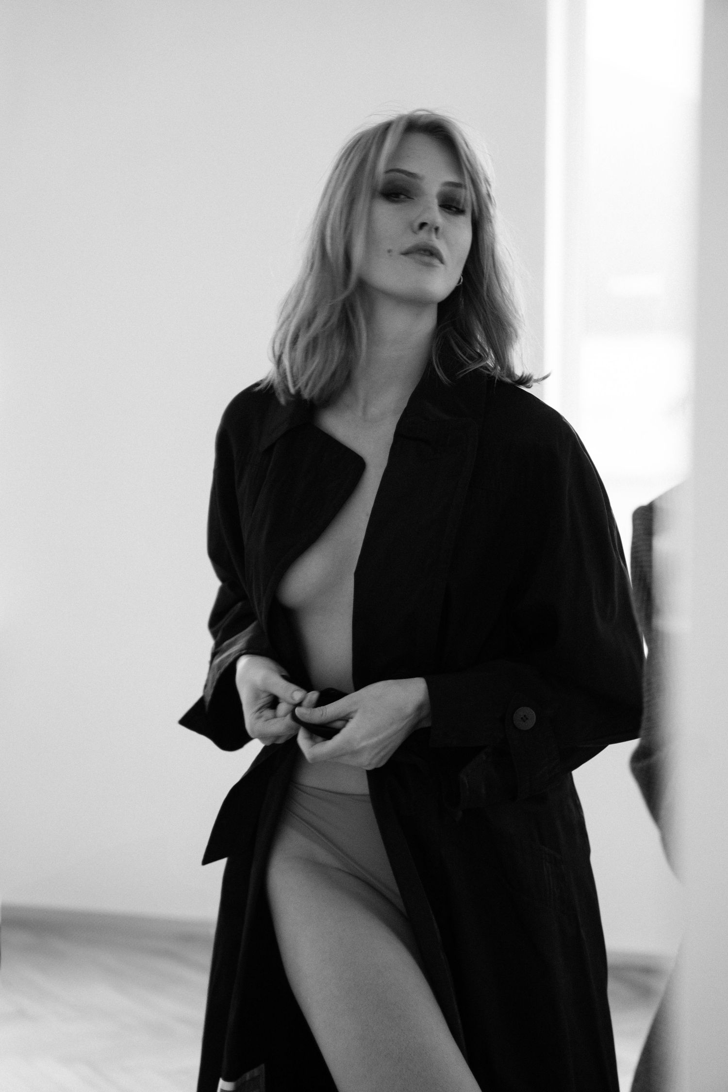 Black and white fashion portrait of woman wearing dark coat in bright room.