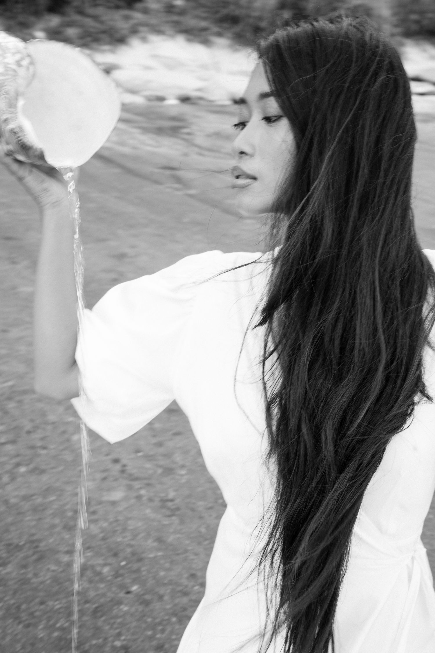 Black and white portrait of woman with long hair pouring water from a sea shell at the ocean