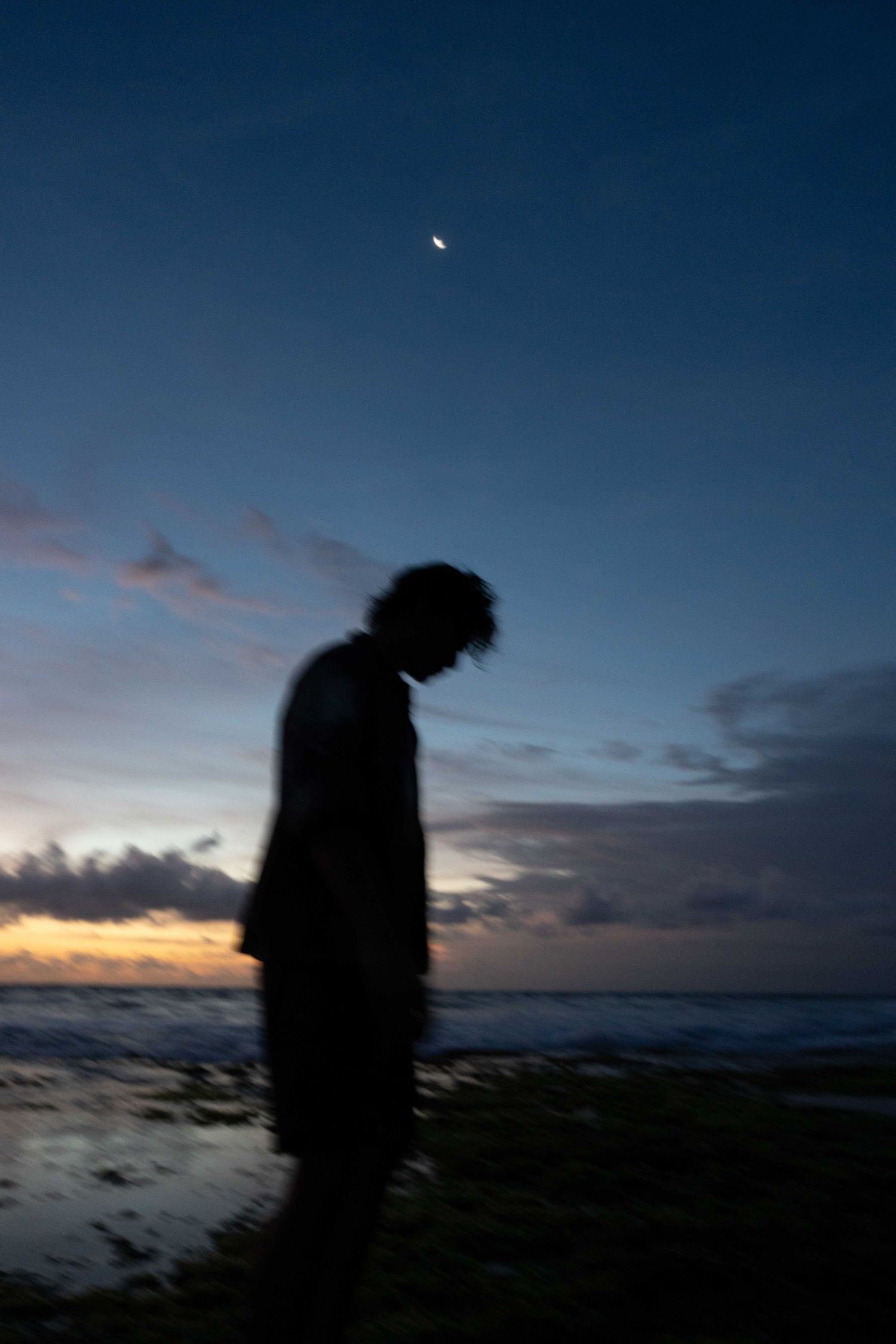 Silhouette of person standing by the ocean at dusk beneath crescent moon.