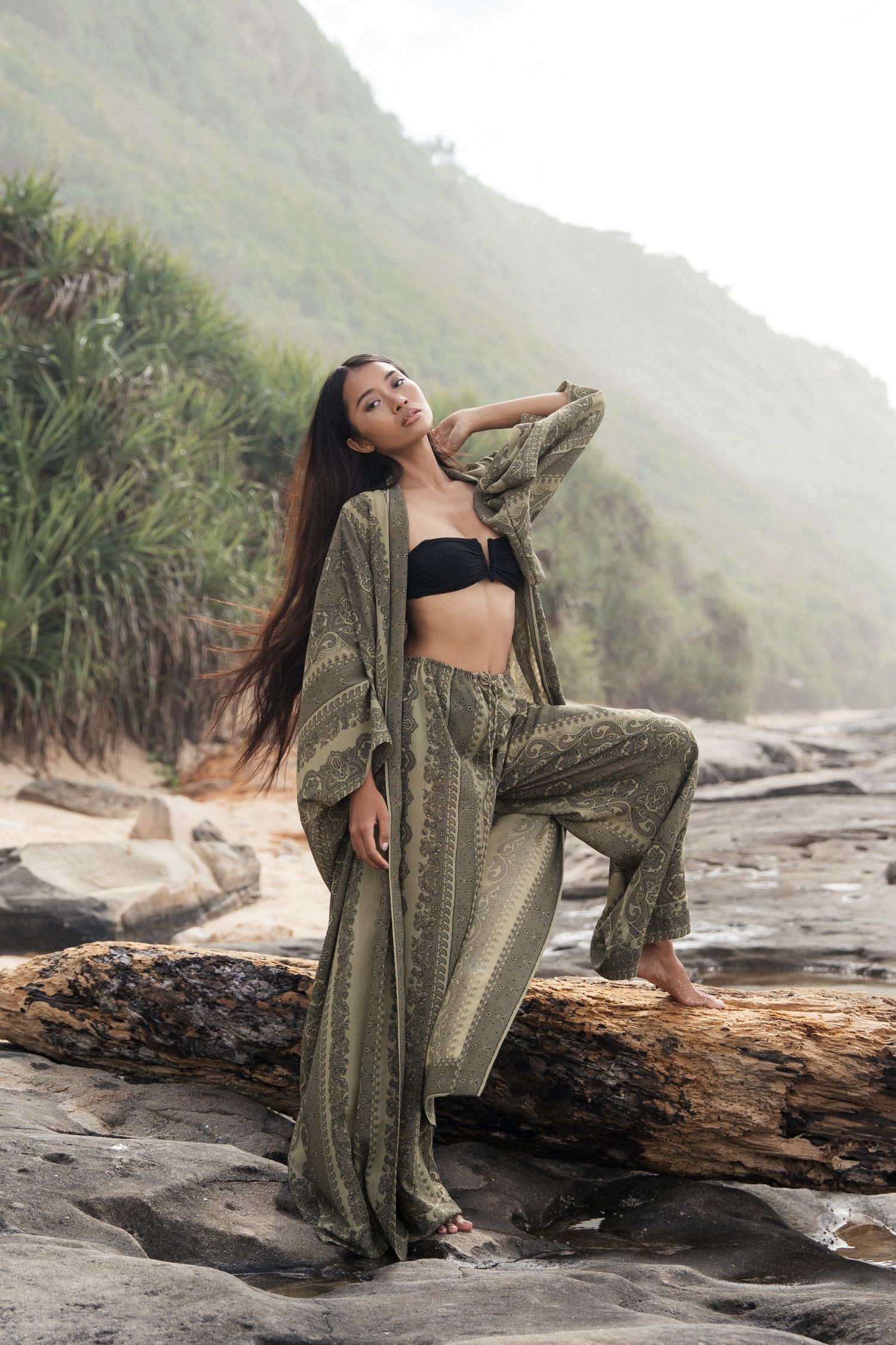 Woman posing on driftwood wearing textured green outfit in misty coastal landscape. Bali, Indonesia