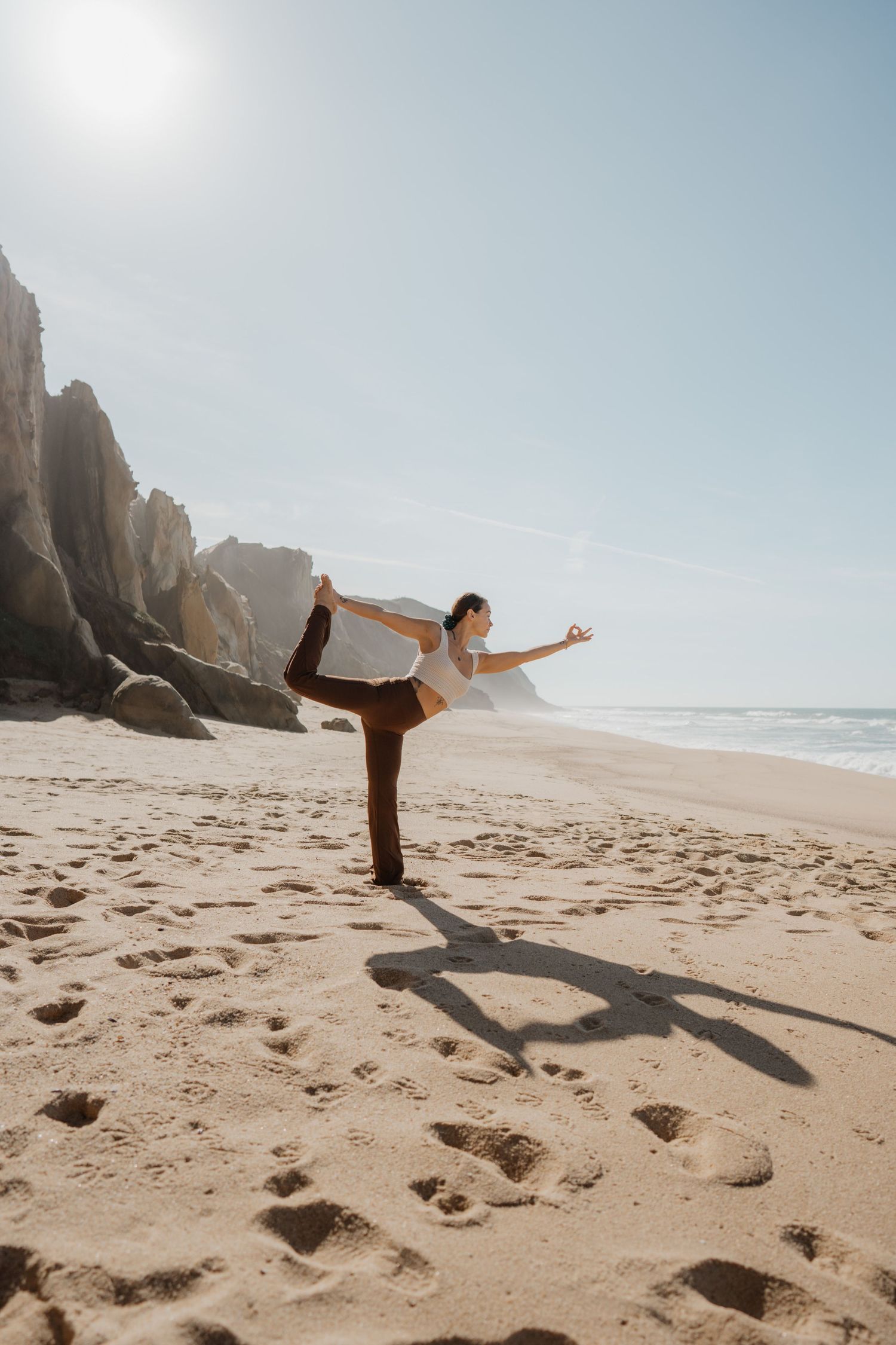 Woman balancing on one leg on sandy beach with ocean in background.