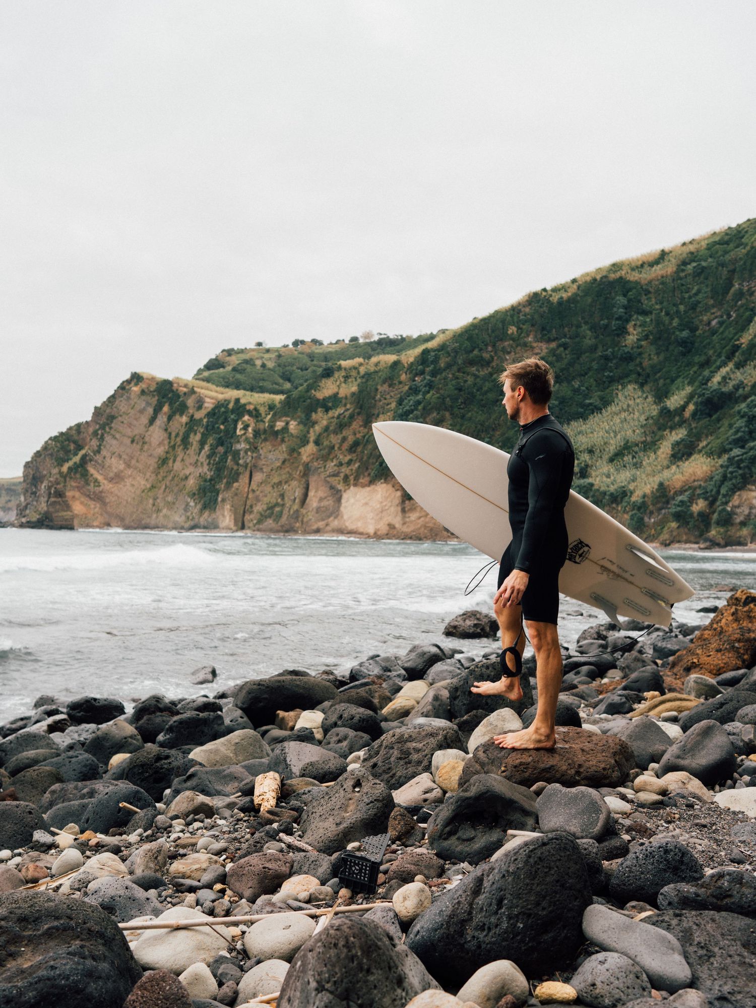 Surfer standing on rocky shoreline holding surfboard and facing the ocean.