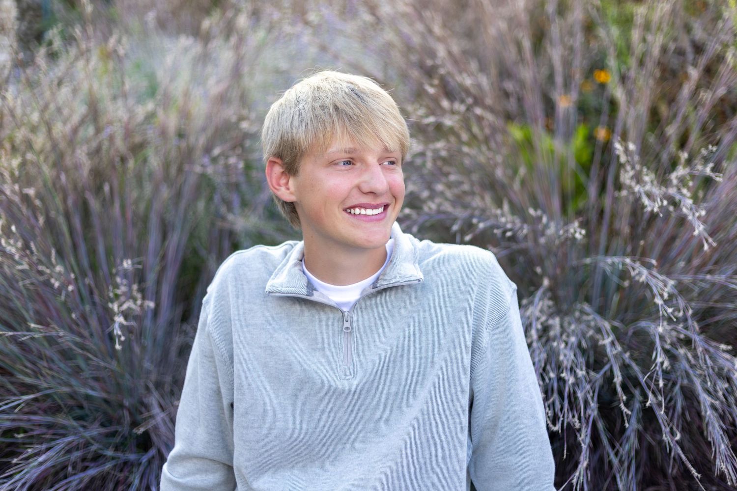 KC portrait photography. Professional senior photo of a young man seated in front of colorful grasses smiling and looking away from camera.