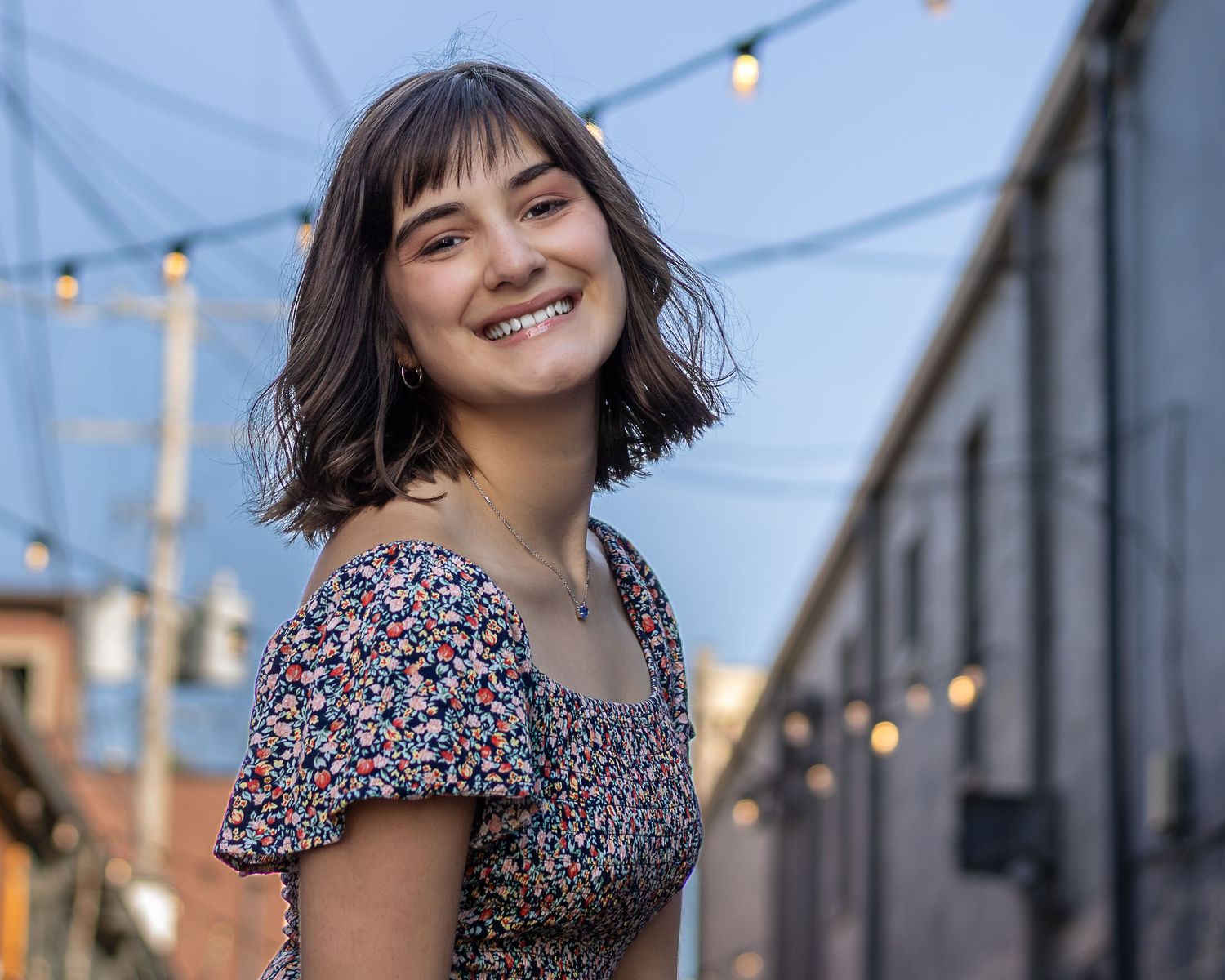 KC portrait photography. Professional senior photo of a young woman twirling and smiling in front of an alley with strung lights.