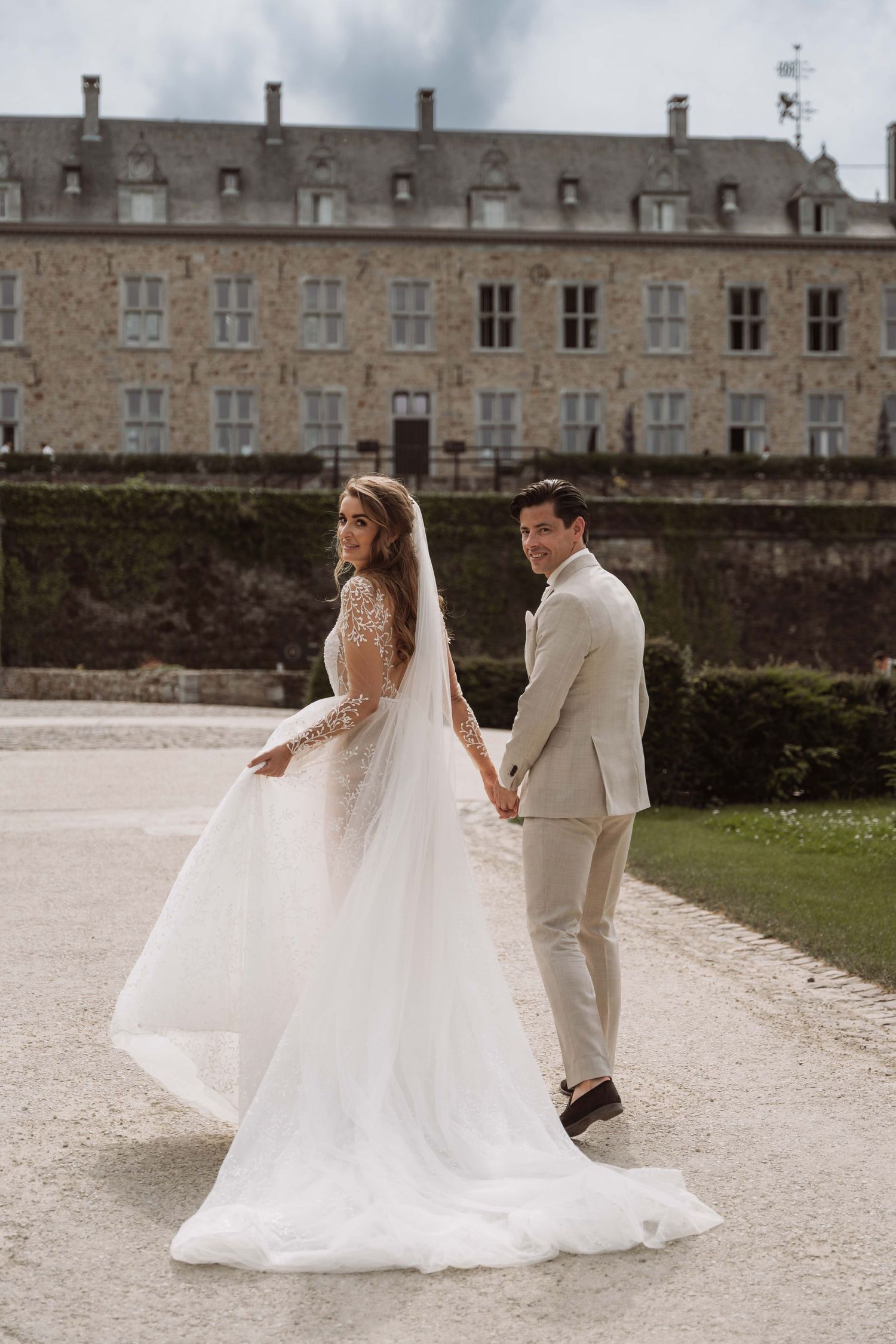 A couple in wedding attire walks together on a stone path outside a historic stone building.