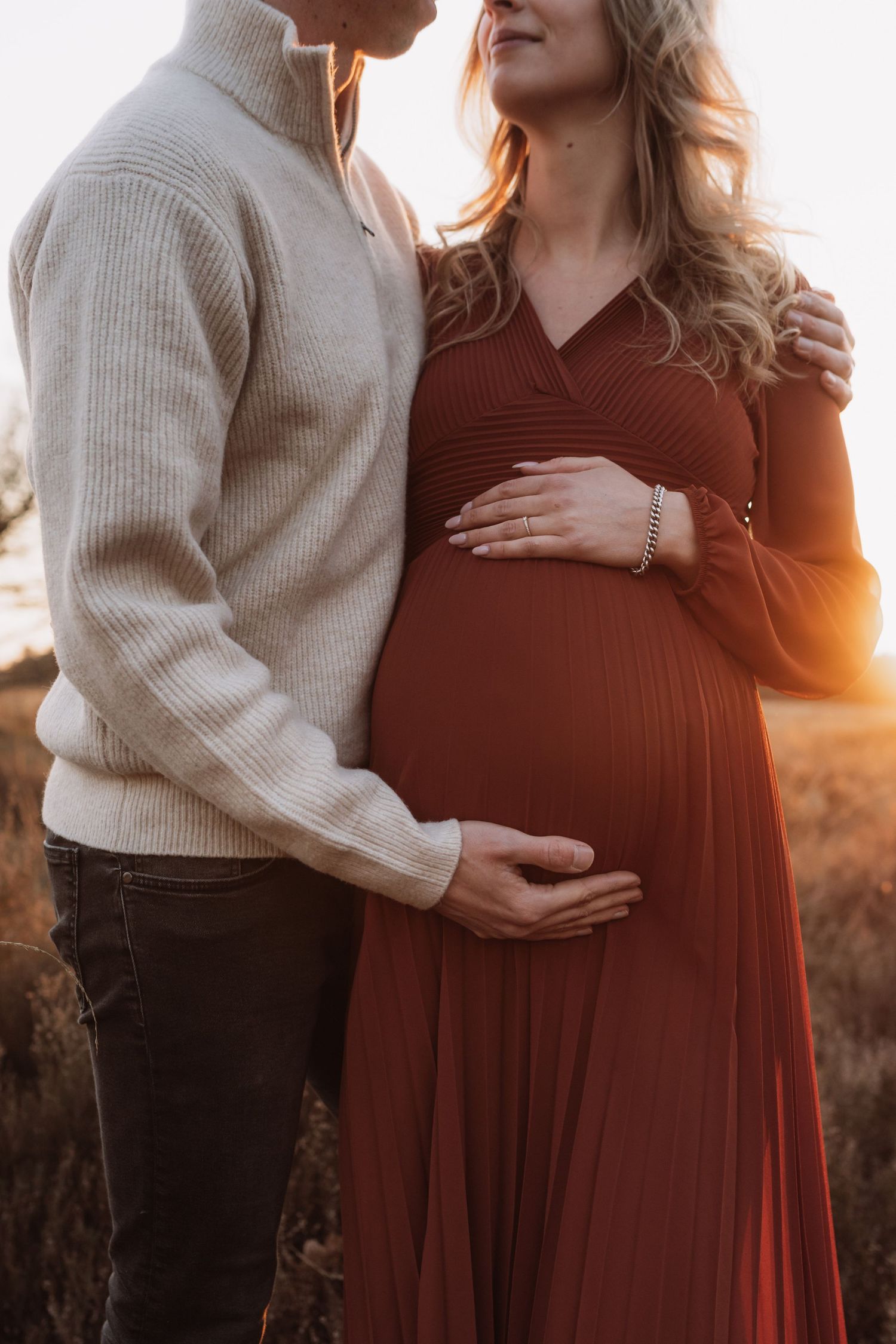 Intimate couple's maternity shoot at golden hour with person in cream sweater and burgundy dress.