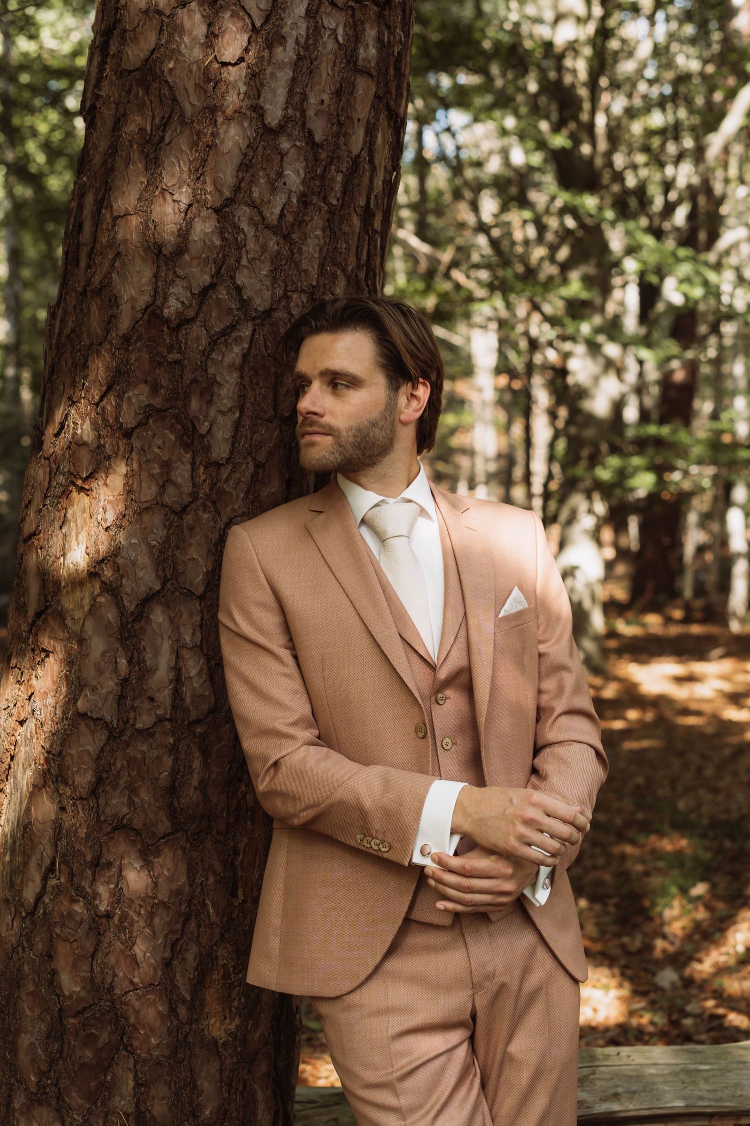 A person in a tan suit and white tie poses thoughtfully against pine trees in a forest setting.