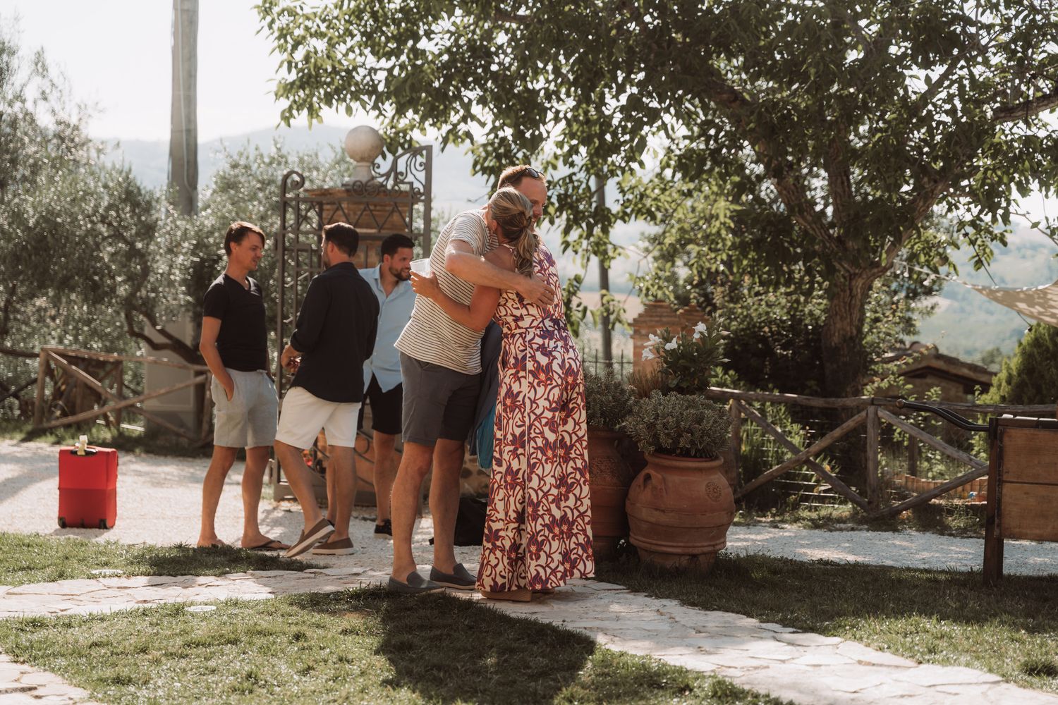 People socialize outdoors on a garden patio surrounded by trees and planters.