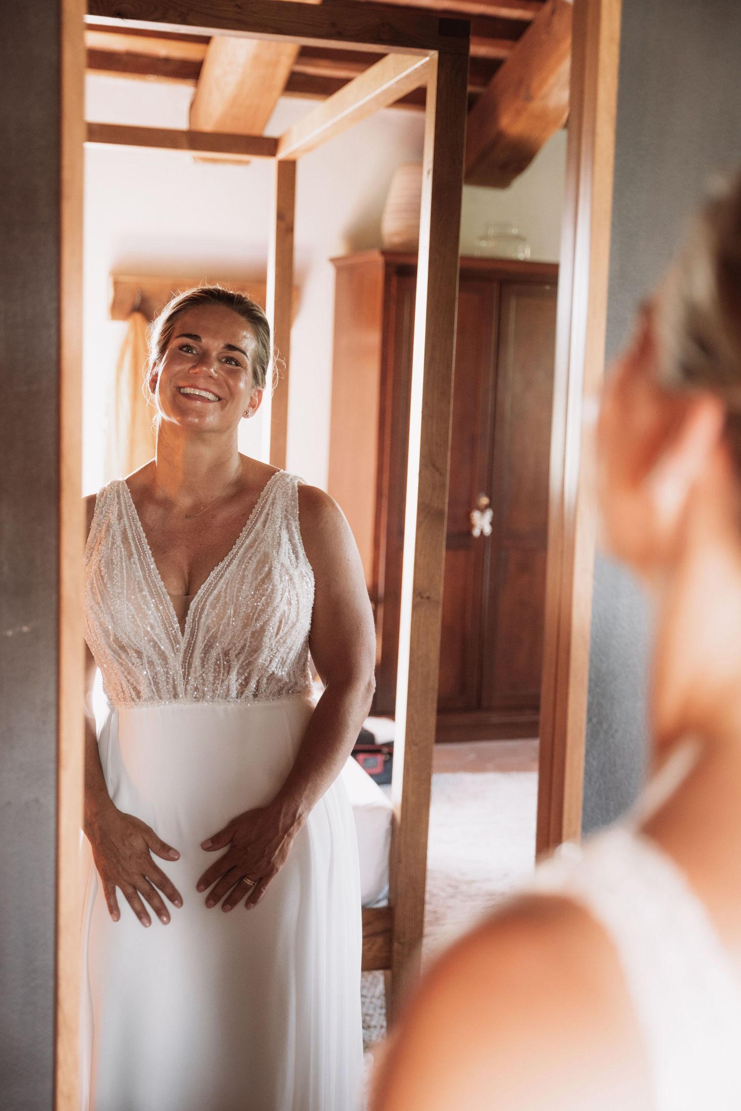 A bride in a white lace wedding dress smiles while getting ready in a sunlit room with wooden beams.