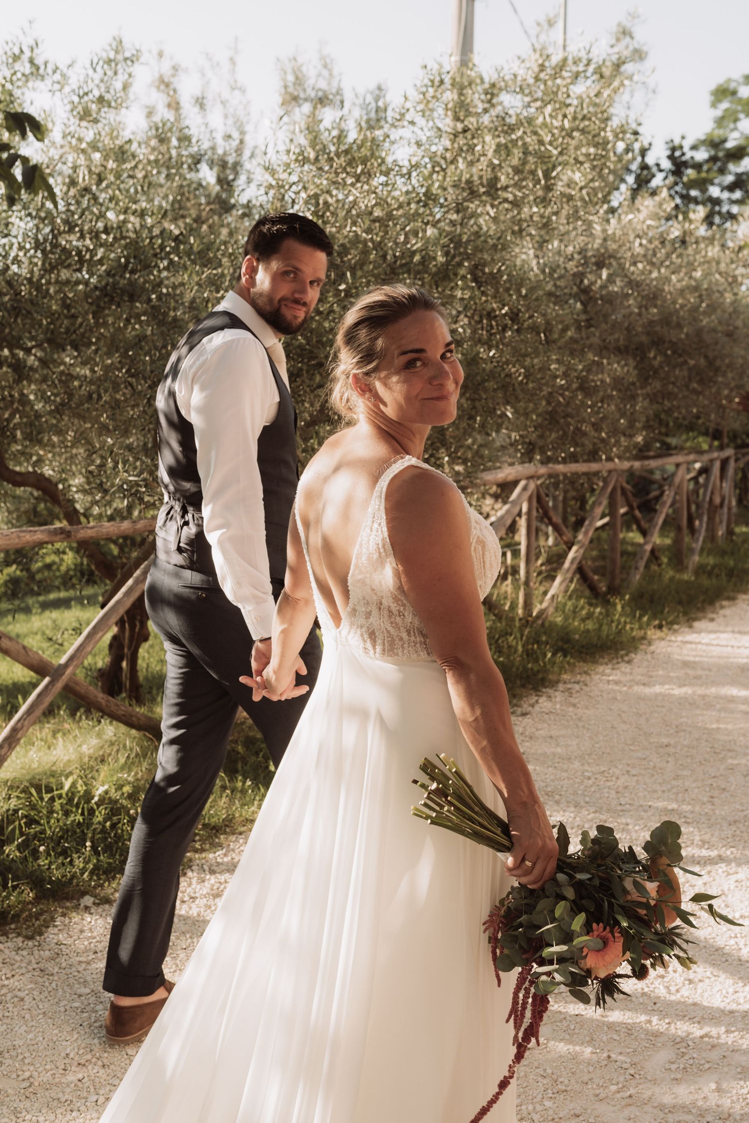 A newlywed couple poses on a rustic path surrounded by olive trees, with the bride holding a bouquet.