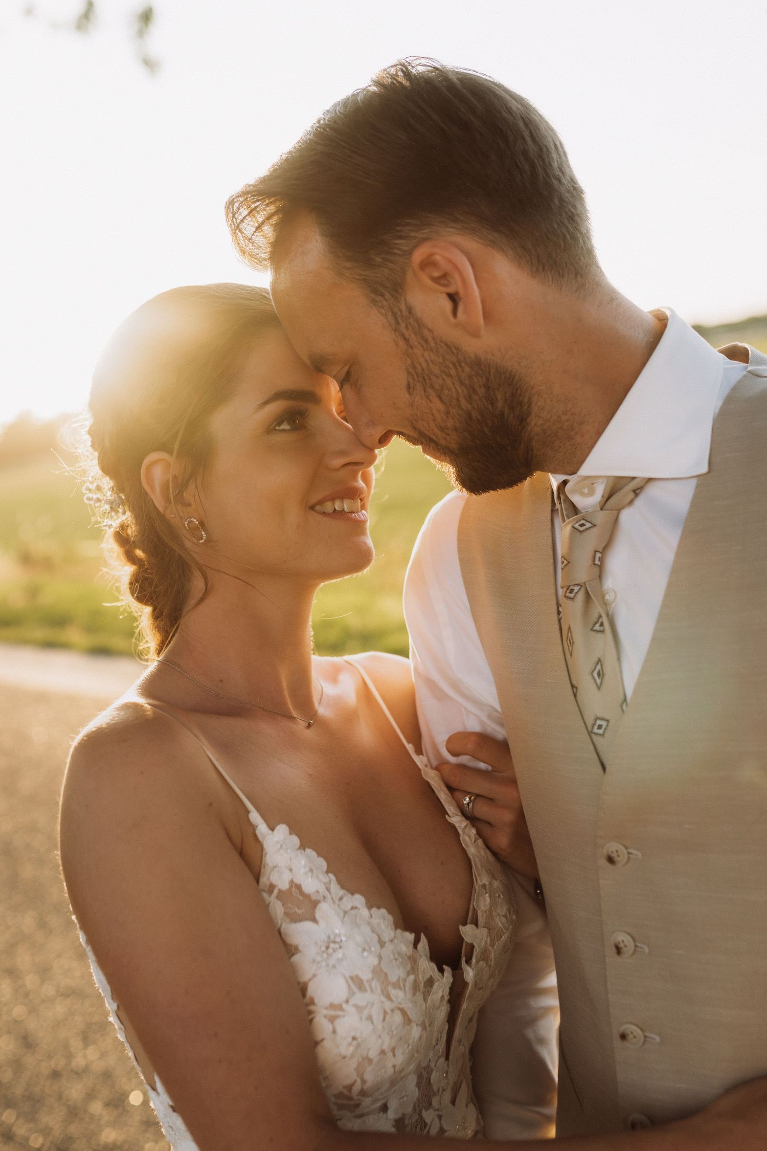 A romantic wedding couple embraces in golden sunlight wearing a white lace gown and beige suit.