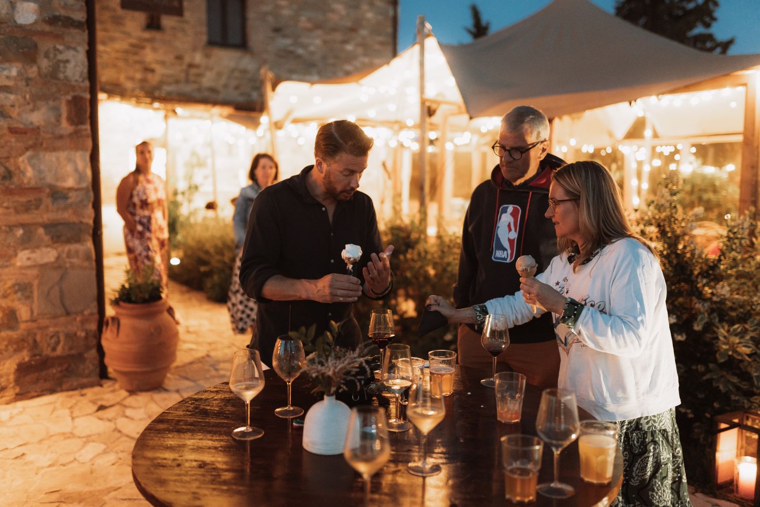 People enjoying drinks and conversation at an outdoor evening event with warm lighting and stone walls.
