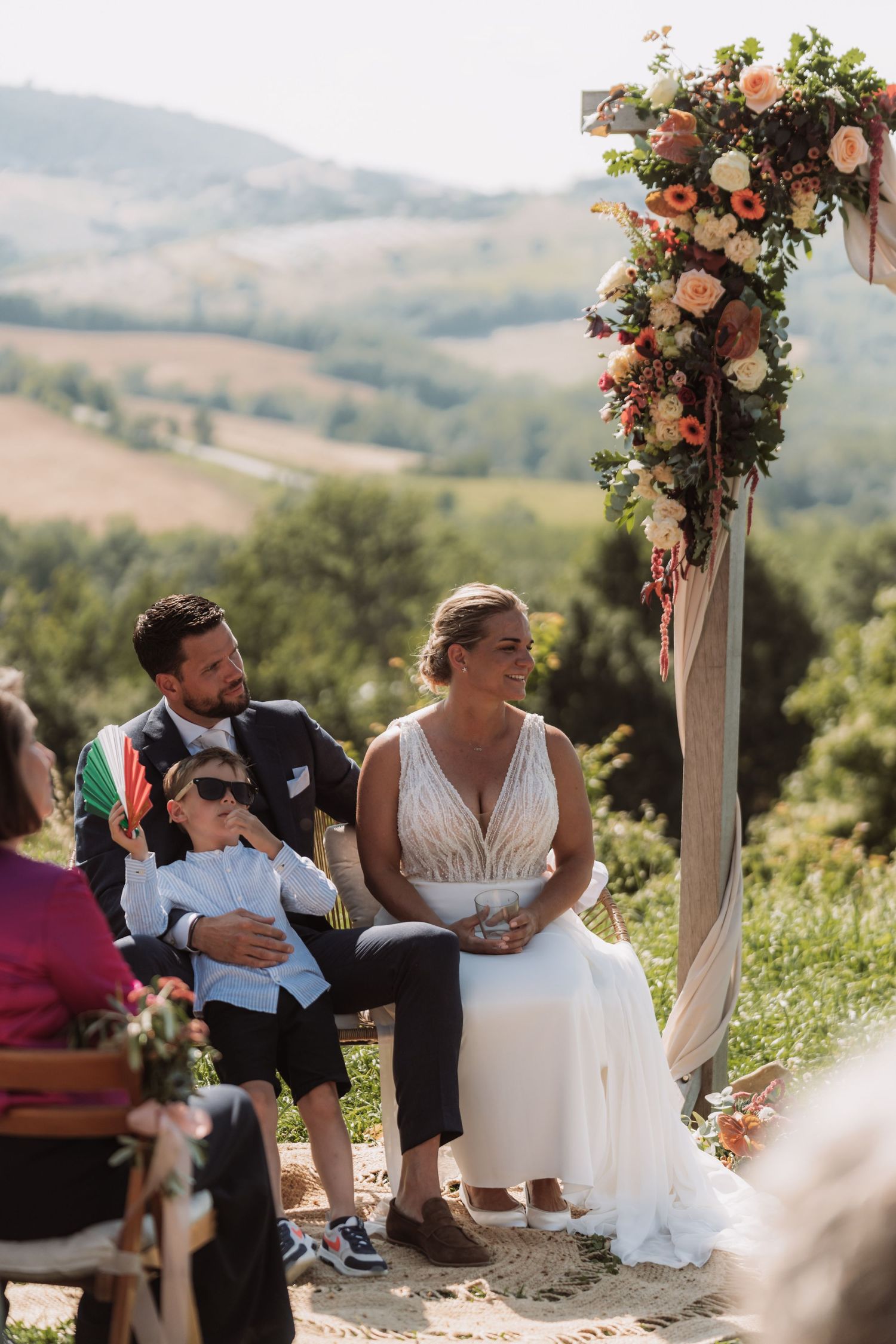 A wedding ceremony takes place outdoors with a floral arch and scenic mountain views in the background.