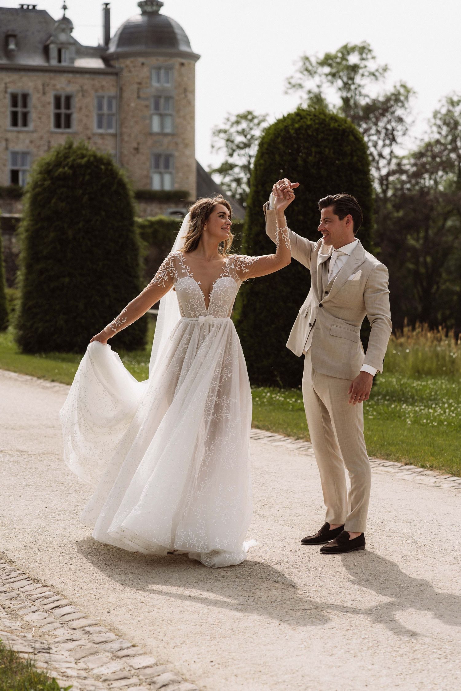 A bride and groom dance playfully together on a gravel path outside a historic stone castle on their wedding day.