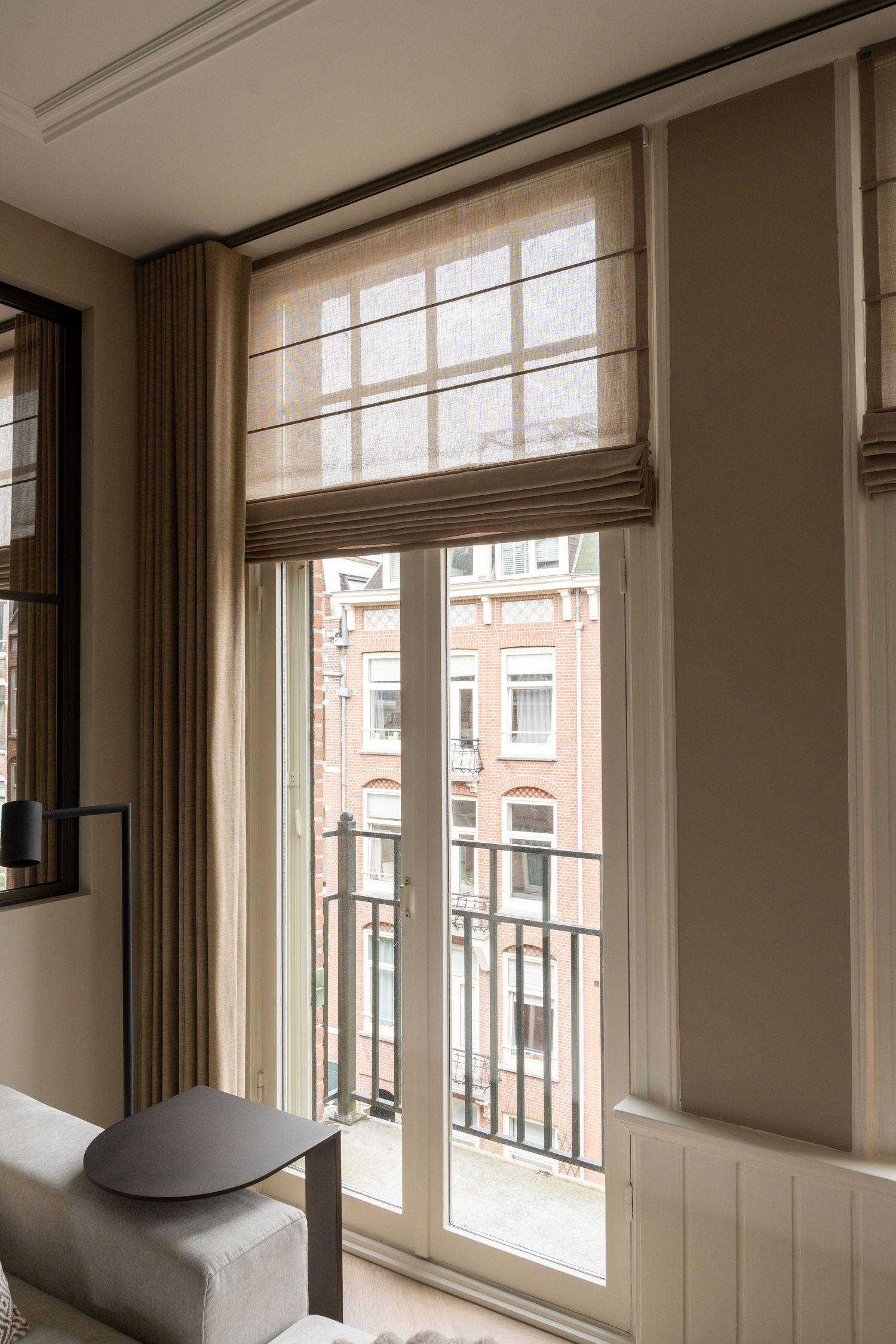 Modern apartment interior with large windows, white walls, and beige curtains showing urban views outside.