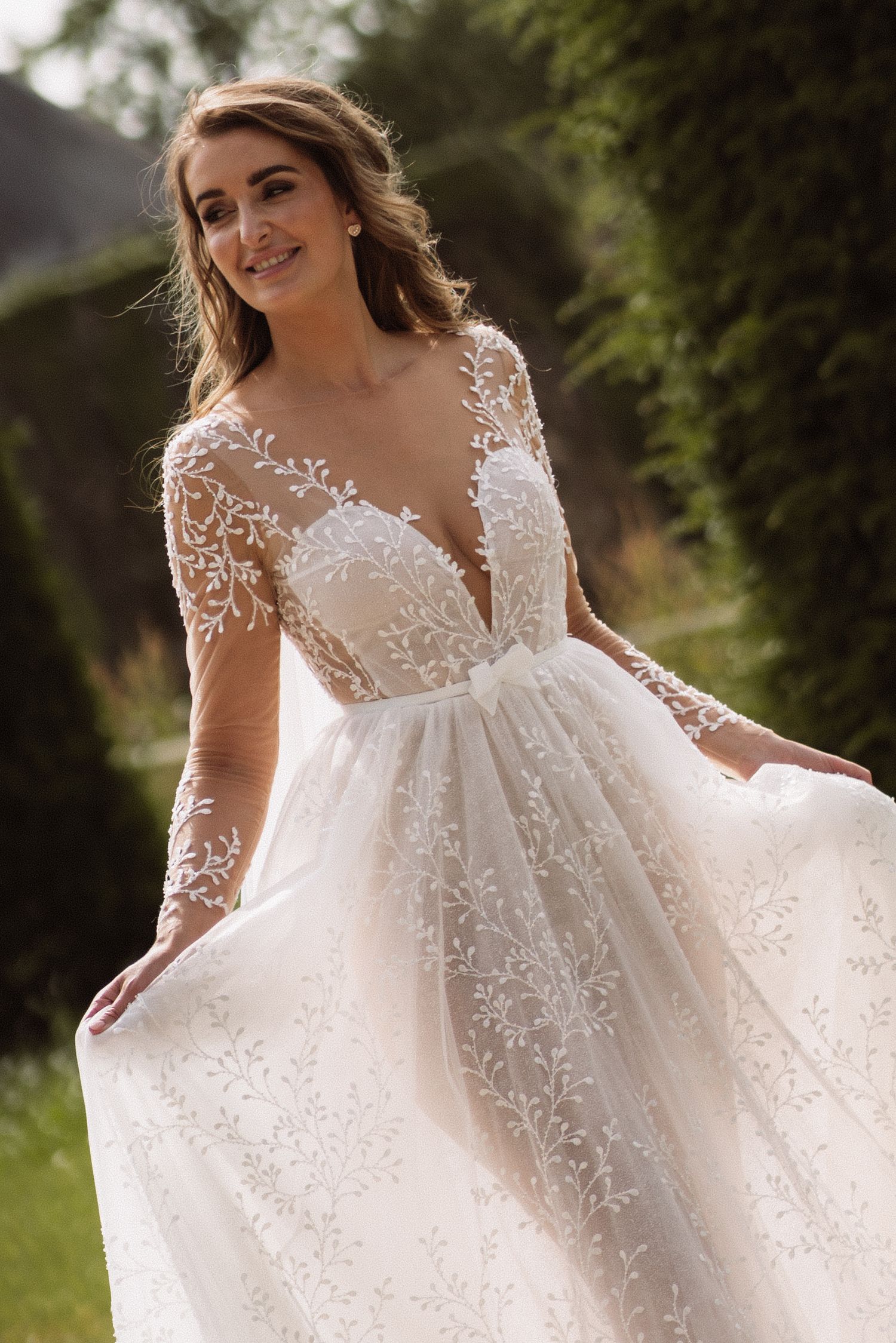 A bride in a flowing lace wedding gown poses in a garden setting.