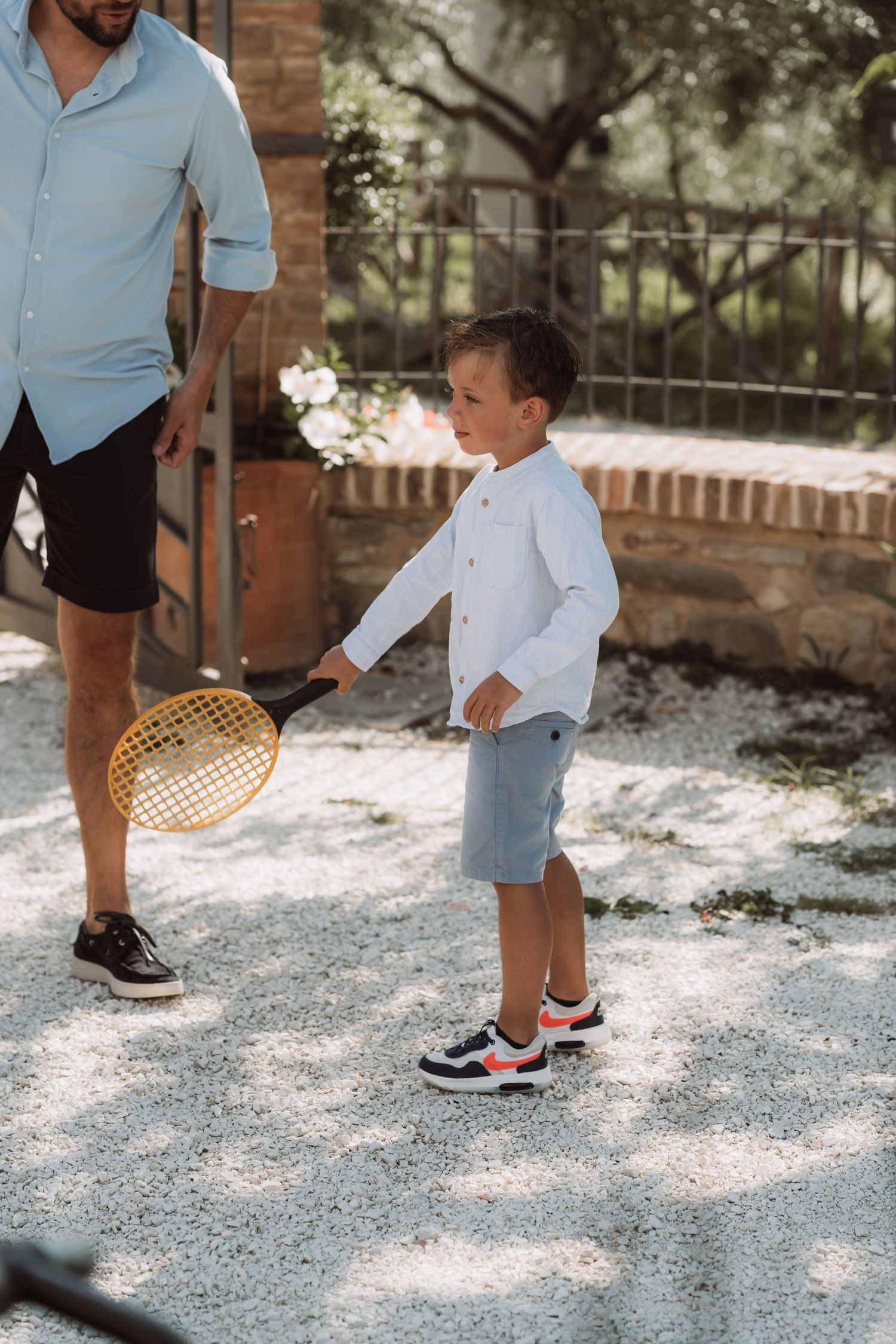 A child in a white shirt and blue shorts holds a tennis racket while standing on a gravel path in dappled sunlight.