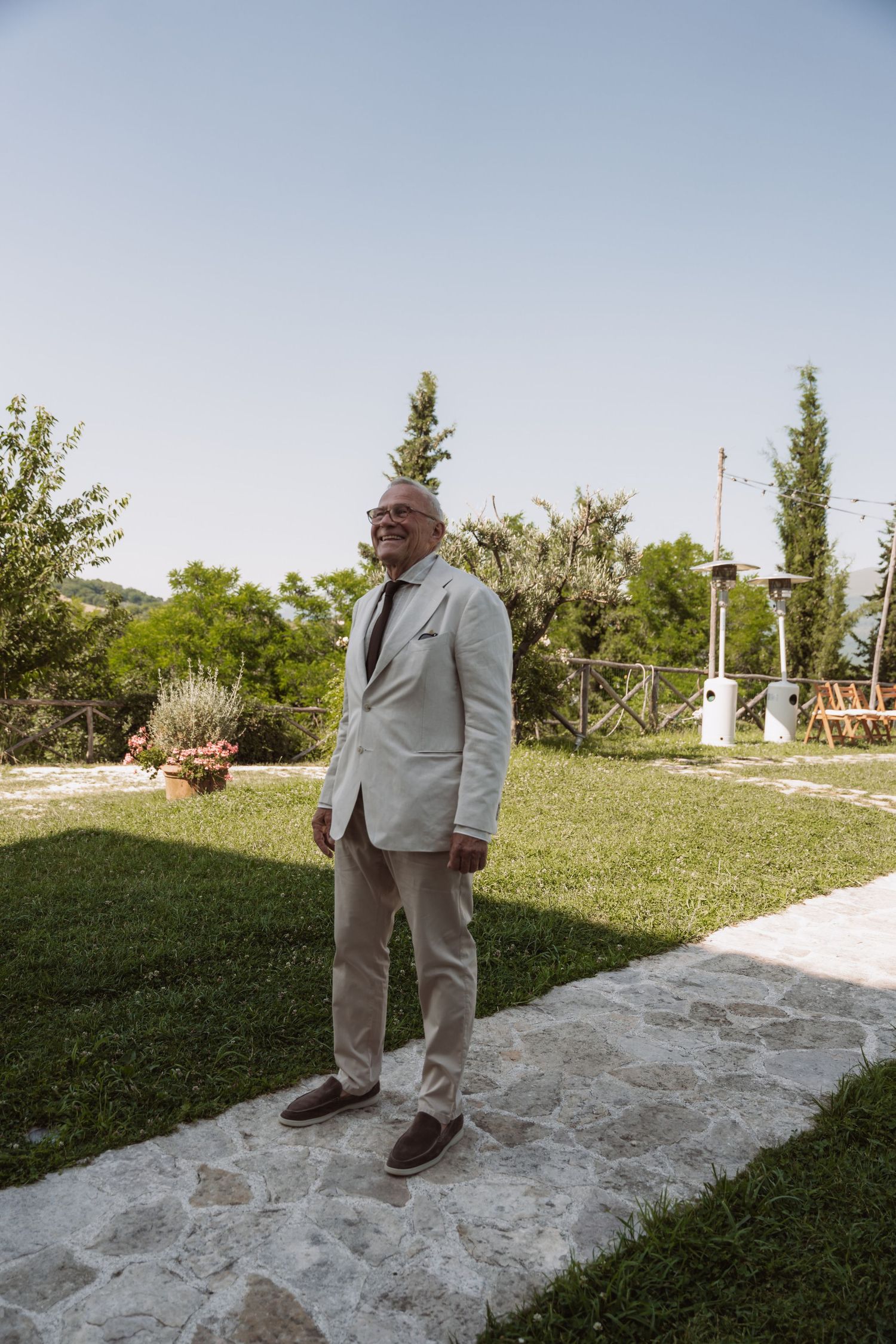 A person in a light-colored suit stands on a white concrete path surrounded by green grass on a sunny day.