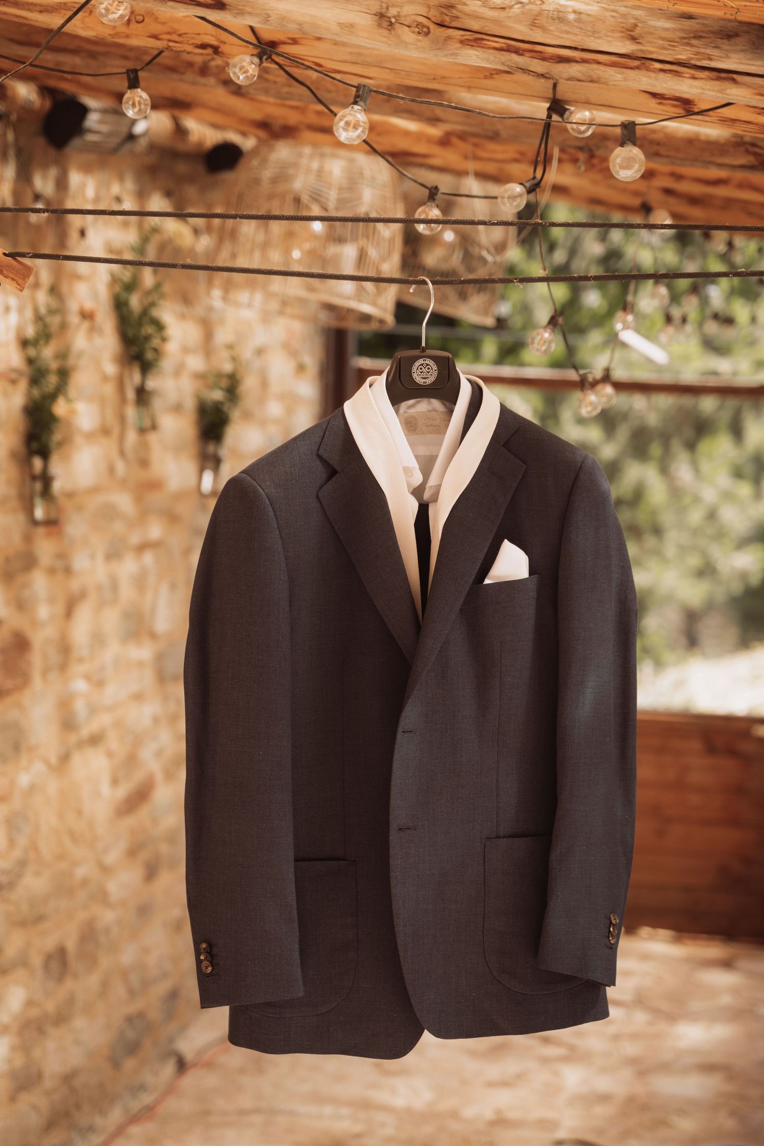 A black suit jacket with white pocket square hangs from wooden beams under rustic string lights.