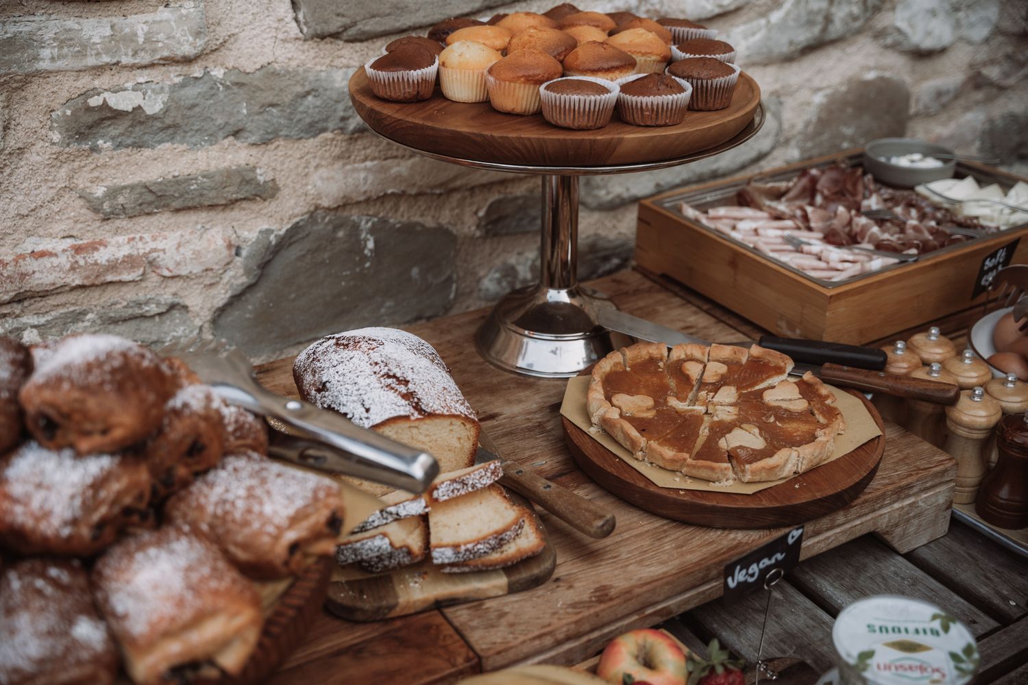 A rustic display of pastries and desserts on wooden boards including macarons, tarts and baked goods on a stone wall backdrop.