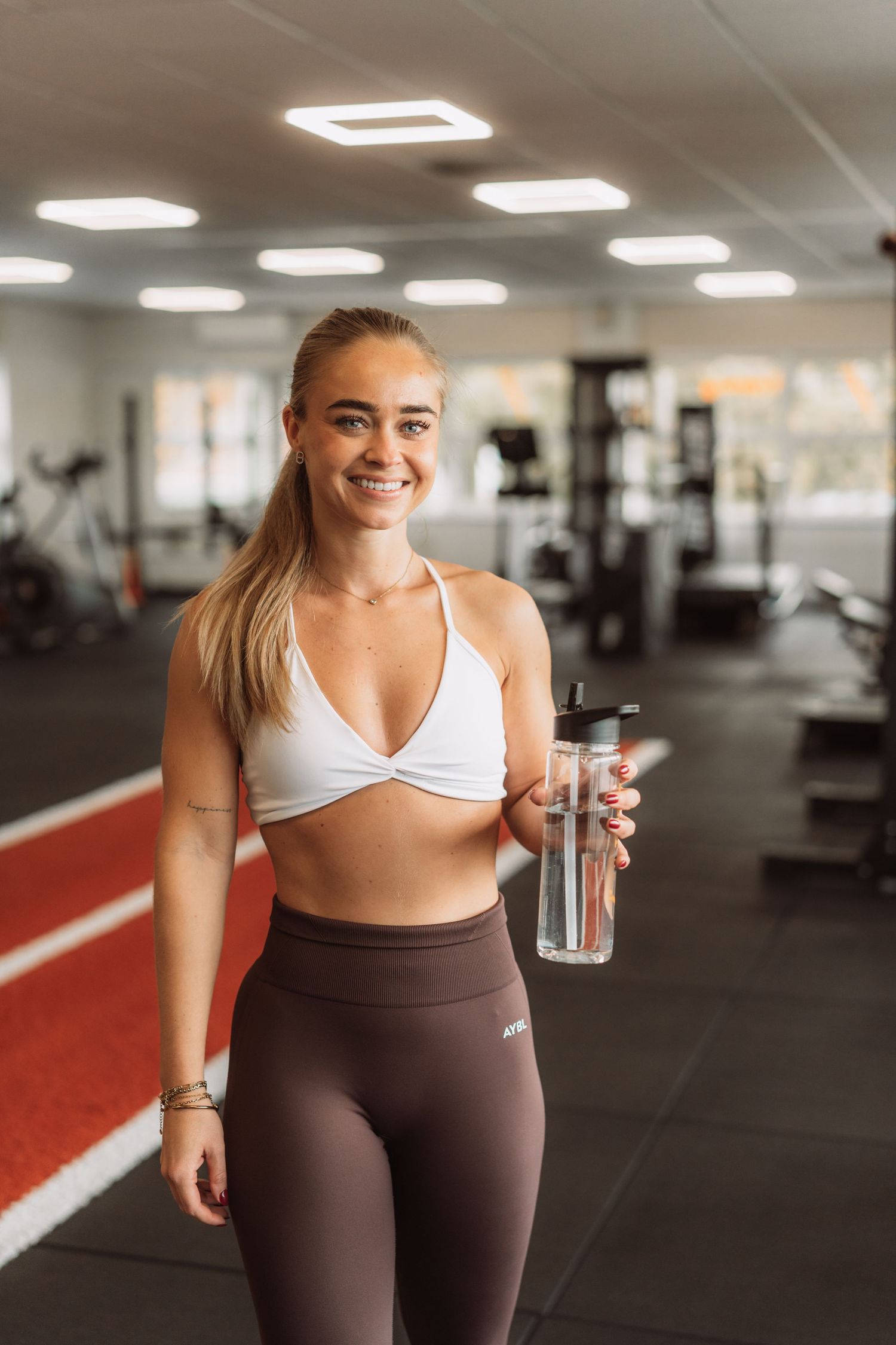 Athletic person in brown leggings and white sports bra holding water bottle during workout at modern gym facility.
