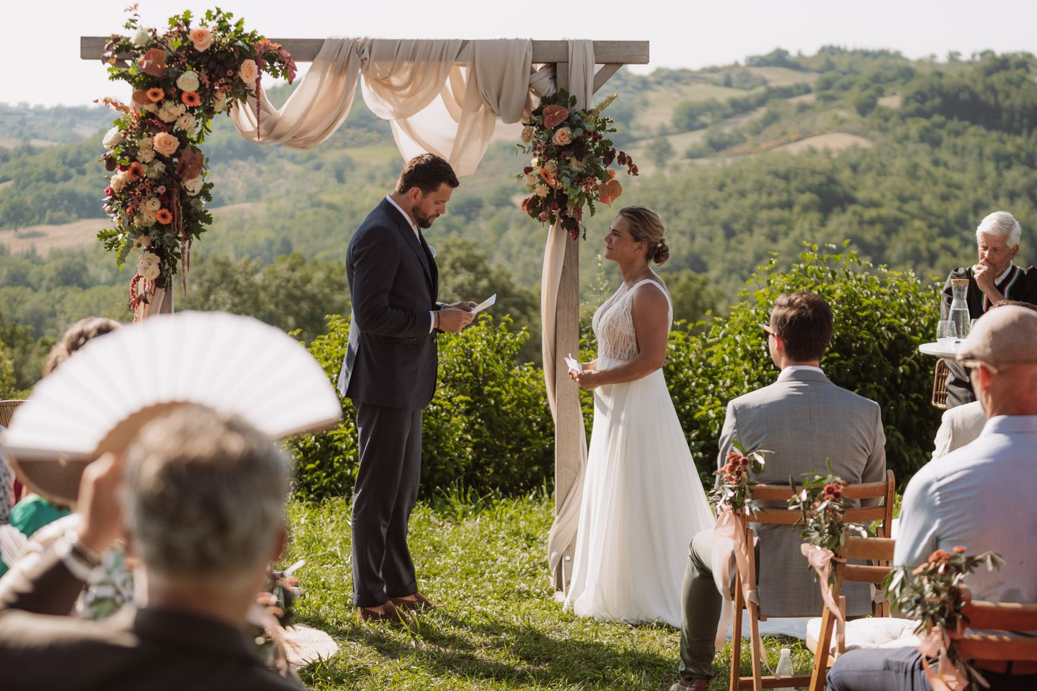 A scenic outdoor wedding ceremony takes place on a hilltop overlooking a lush green valley in Tuscany.
