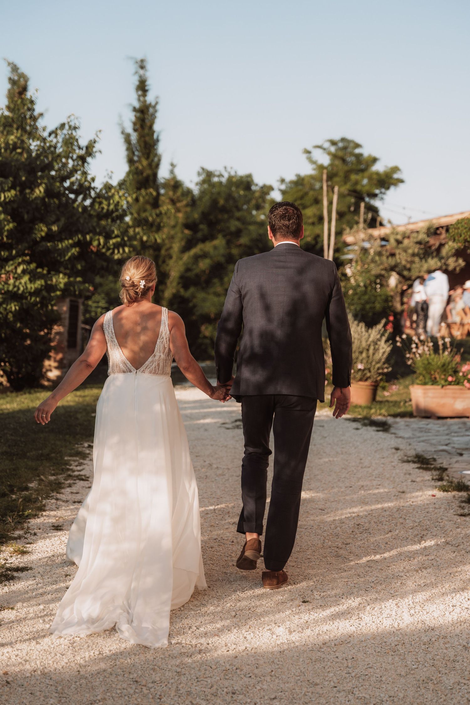 A couple in wedding attire walk hand in hand down a gravel path surrounded by lush greenery on a sunny day.