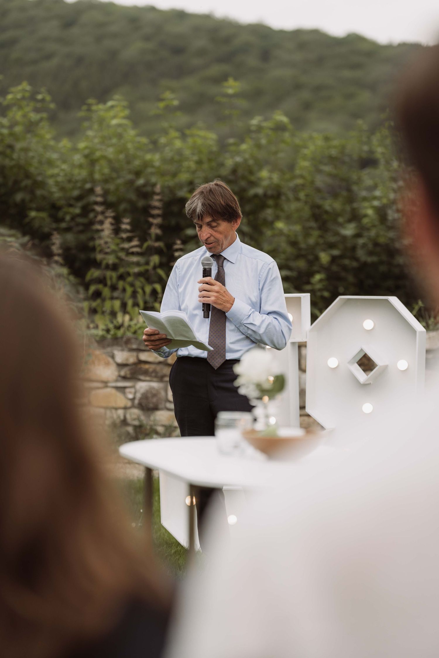 A person in formal attire speaks into a microphone during an outdoor wedding ceremony.