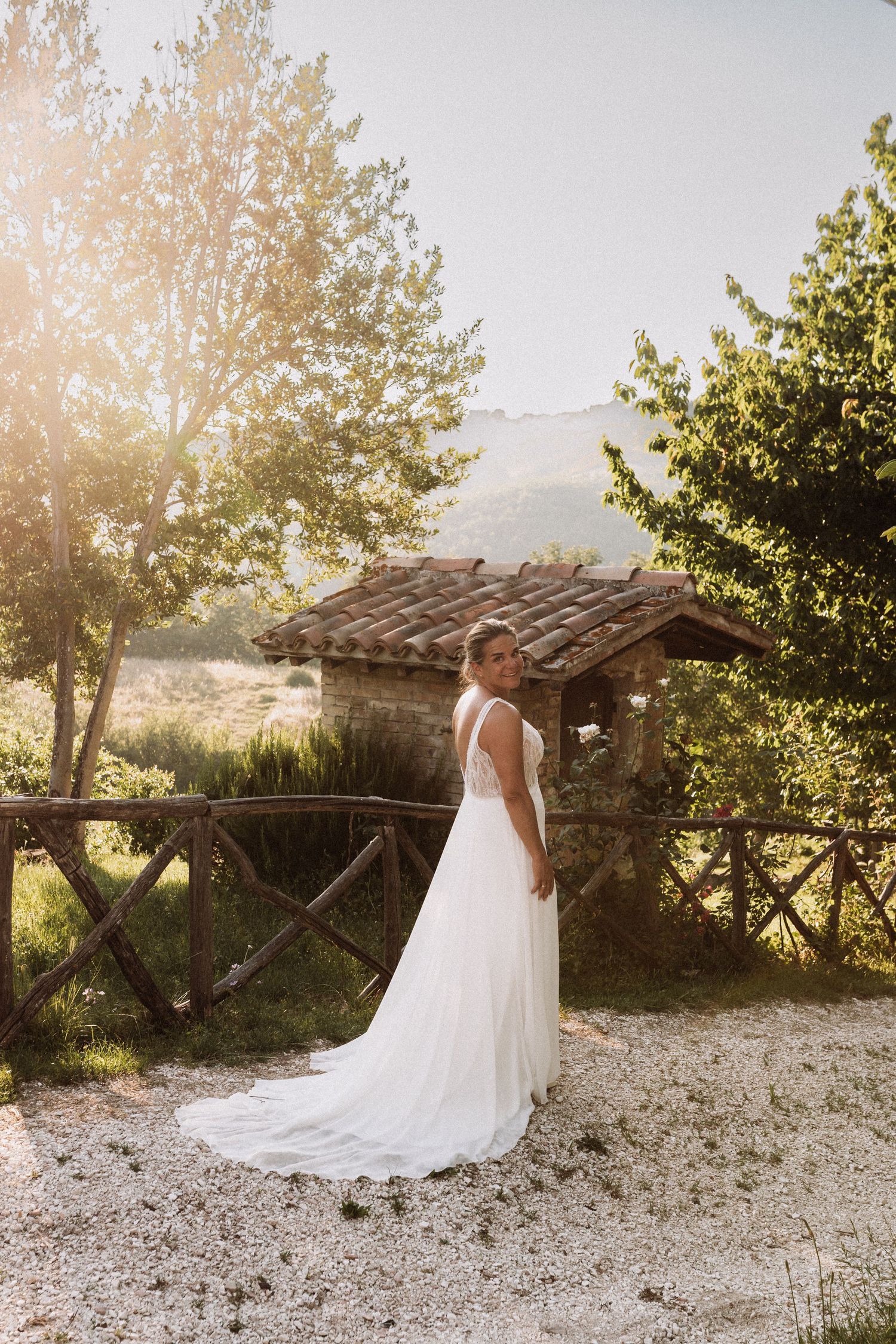 A bride in a flowing white wedding gown stands near a rustic stone structure in golden sunlight at sunset.