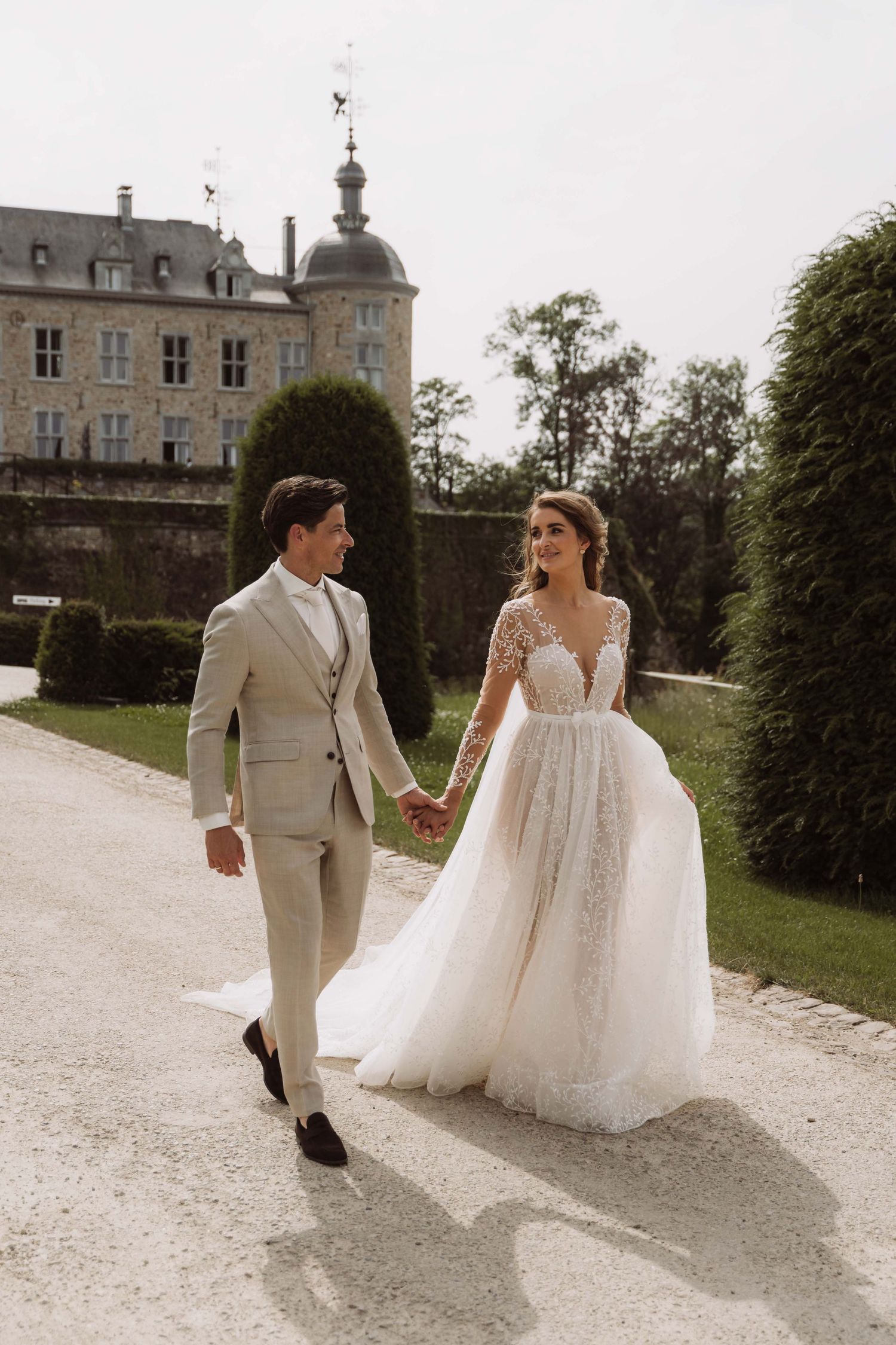 A romantic wedding couple walks along a gravel path beside a grand chateau in France.