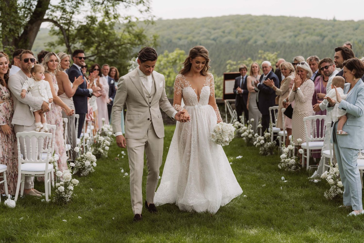 A romantic outdoor wedding ceremony takes place on a lush hillside with guests seated on white chairs and flower petals scattered about.
