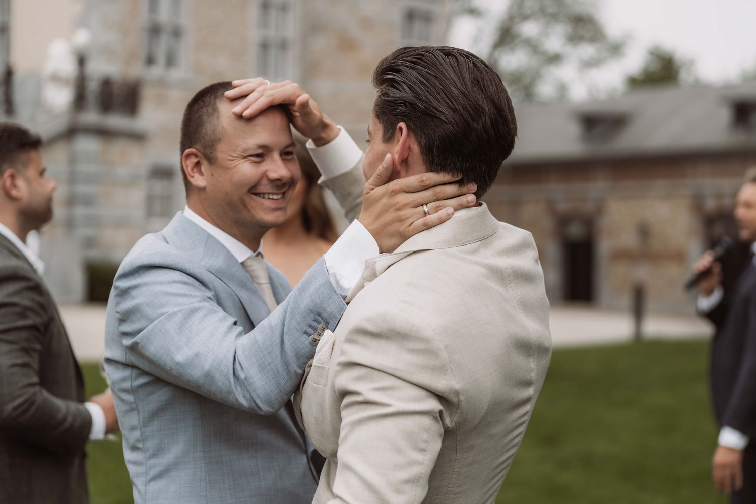 A couple shares a joyful moment during their outdoor wedding celebration on a grassy lawn near stone buildings.