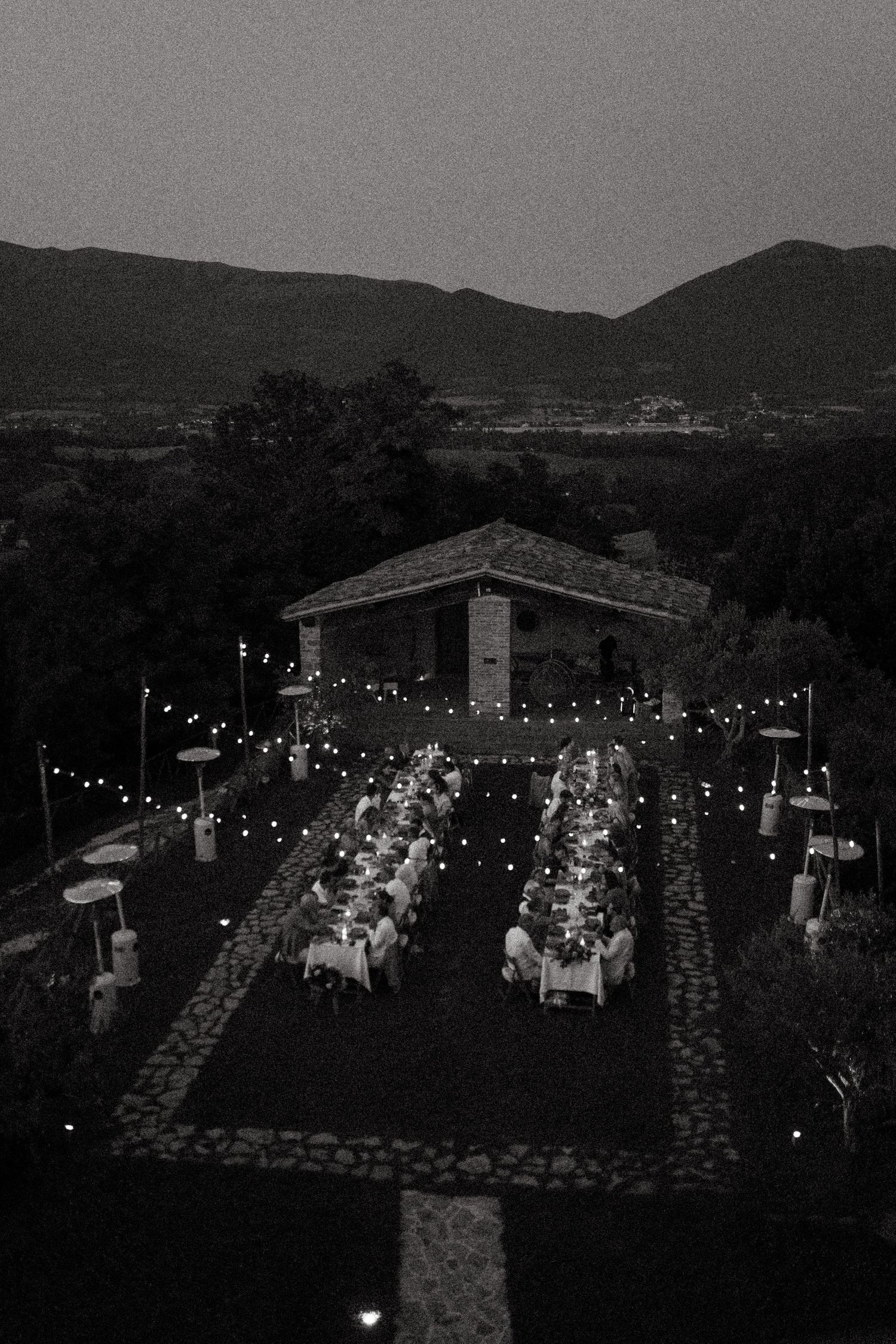 An outdoor evening wedding reception with string lights illuminating tables and guests under the darkening sky.