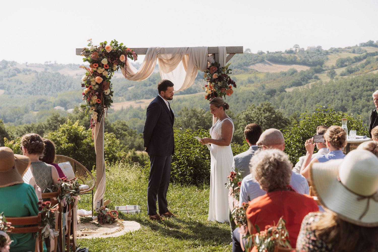 Outdoor wedding ceremony in a scenic hilltop setting with floral arch decorations and guests seated on white chairs.