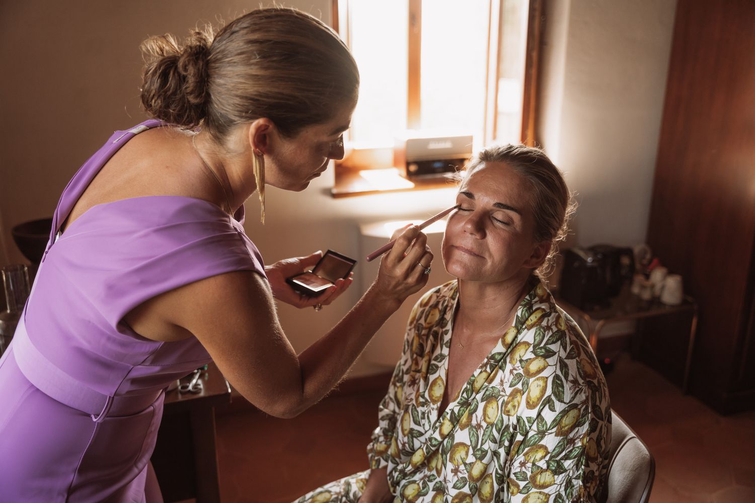 A caregiver in purple attends to an elderly person in a floral dress while sitting in a sunlit room.