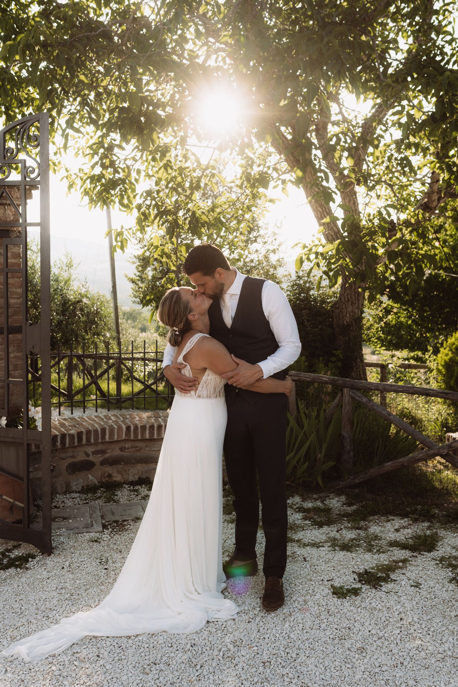A romantic wedding couple embraces in a sun-dappled garden setting with beautiful backlit golden hour lighting.