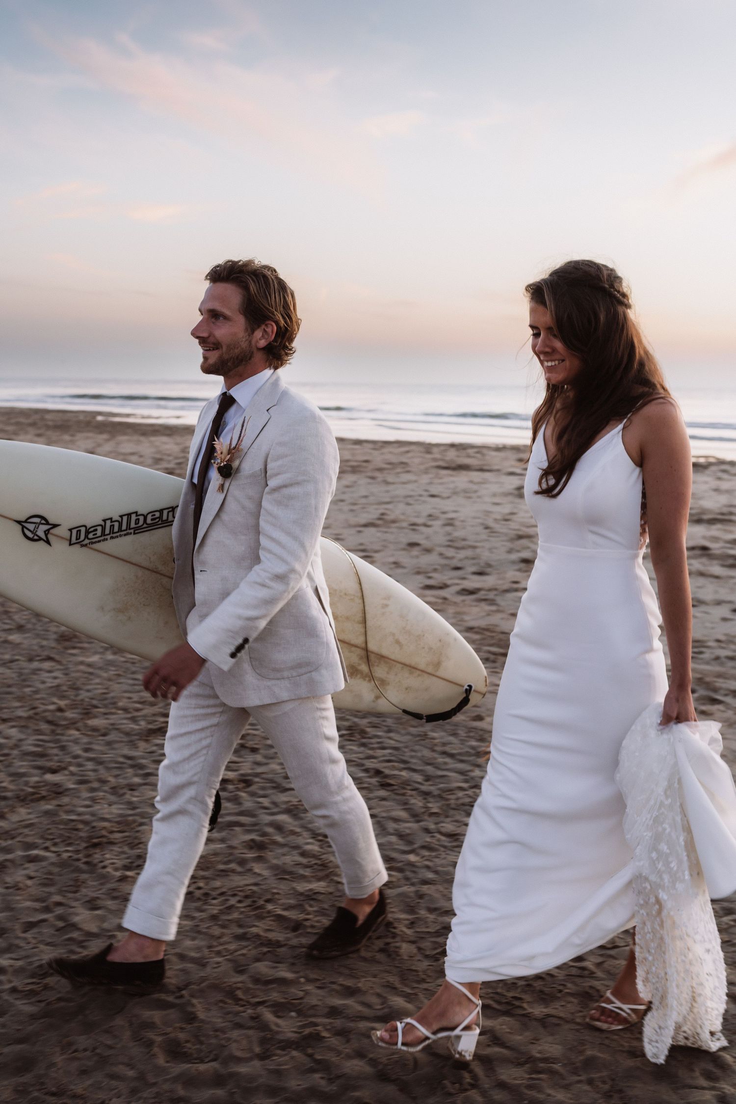 A couple in white outfits walking on a beach carrying a surfboard at sunset.