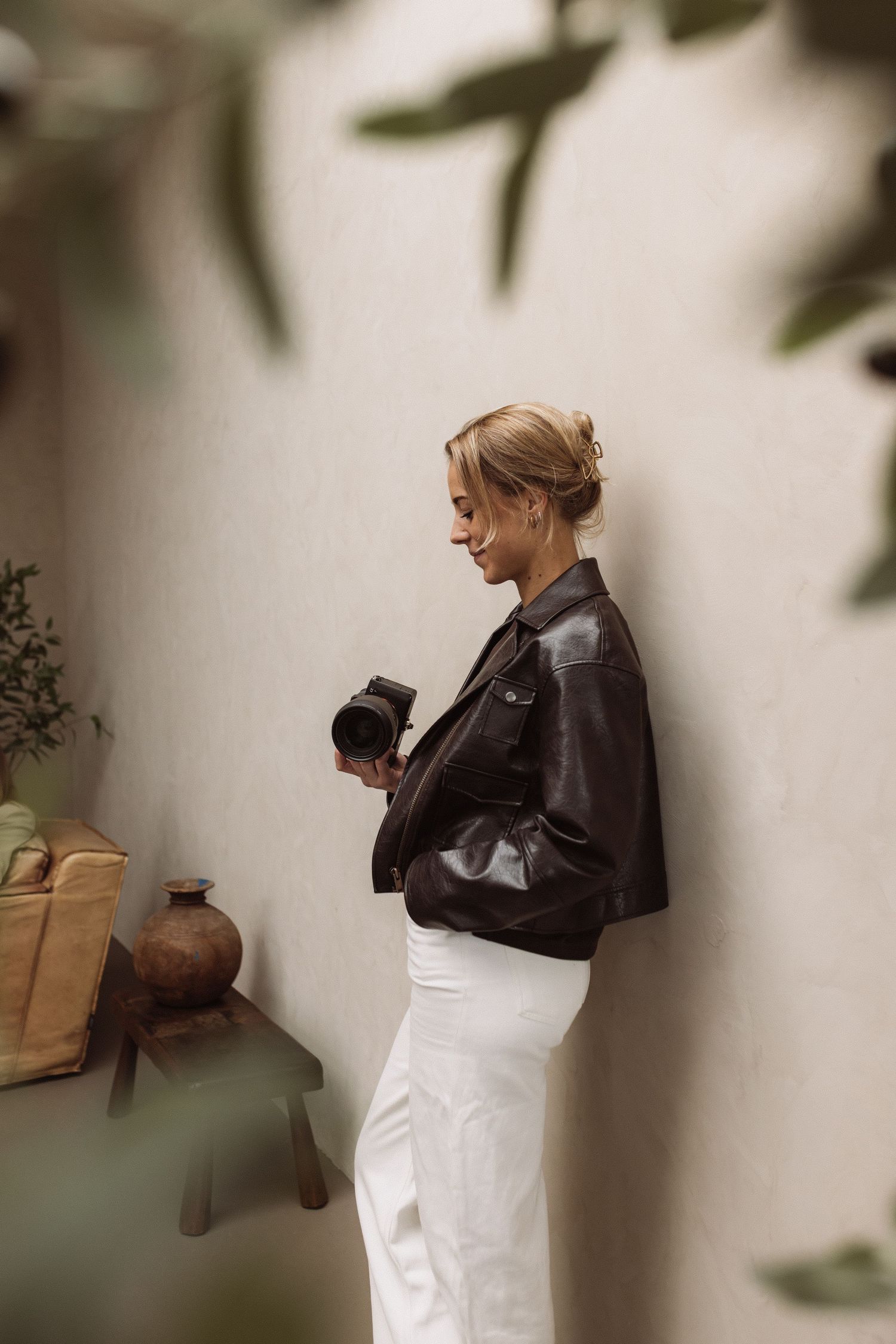 A person in a black leather jacket and white pants stands against a white wall holding a camera.