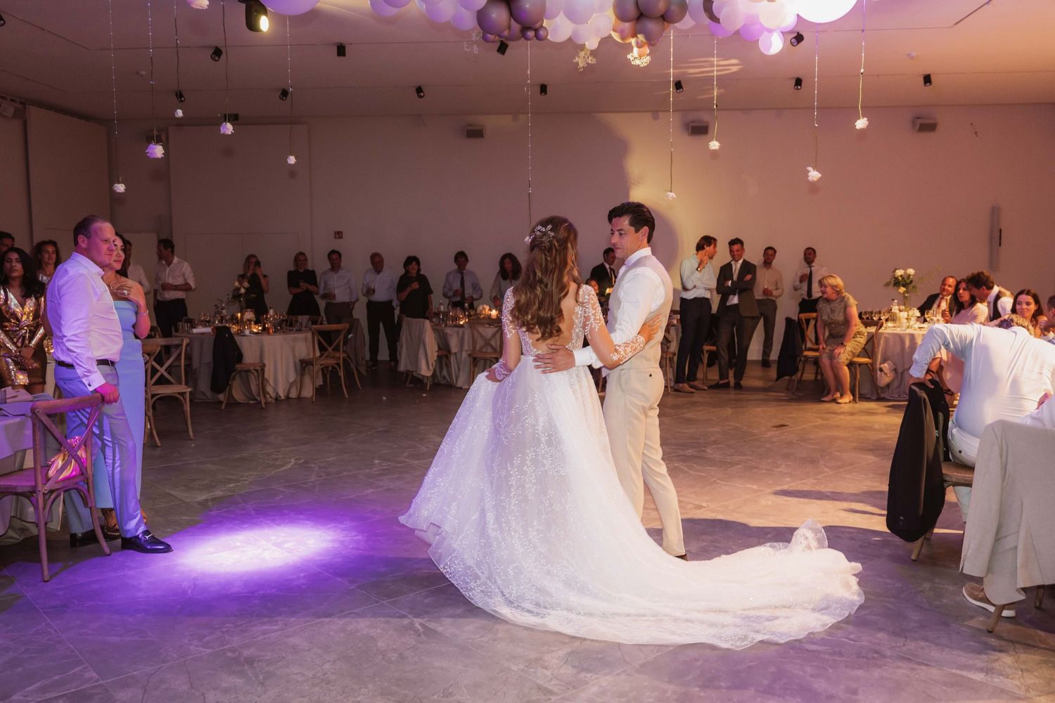 A wedding couple shares their first dance on a white dance floor illuminated by purple lighting in a reception hall.