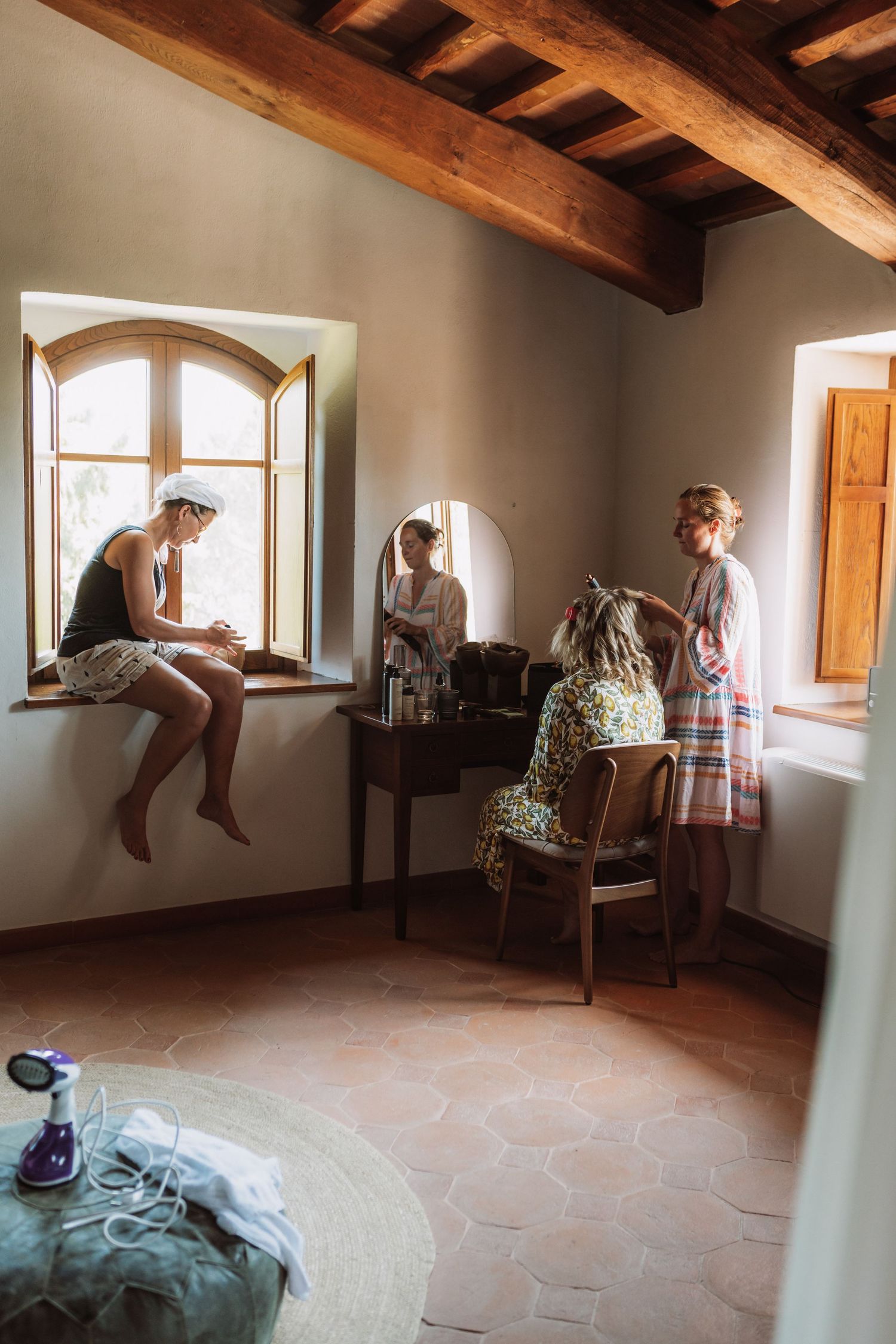 People sitting and standing by rustic windows in a room with exposed wooden beams and stone tile flooring.