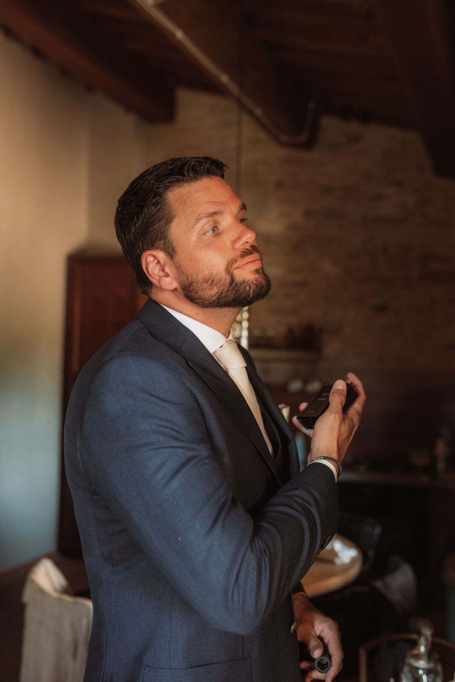 A person in a navy suit adjusts their tie while looking upward in a dimly lit rustic room.
