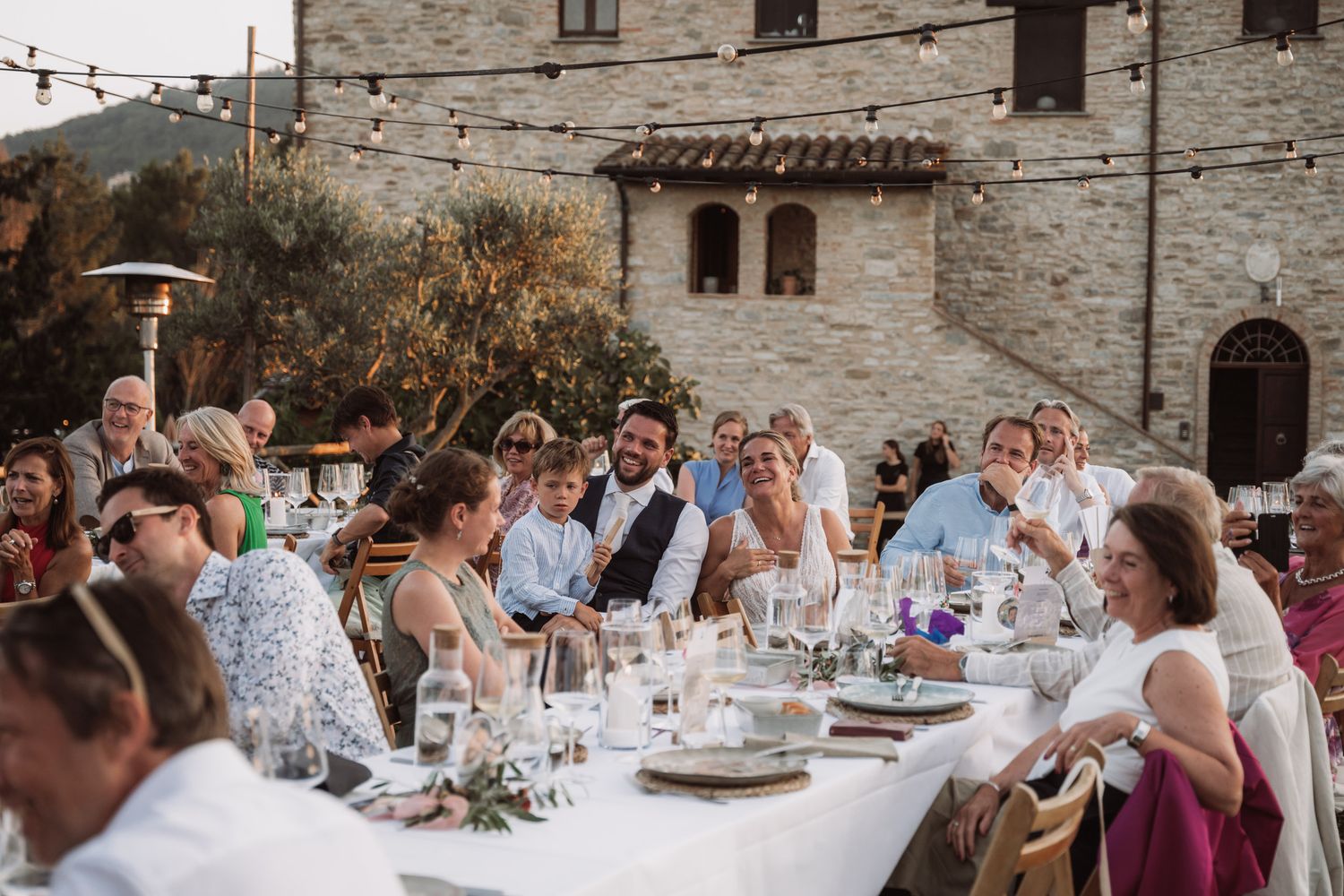 A panoramic view of an outdoor wedding reception with guests dining at long tables in a rustic Italian courtyard setting.
