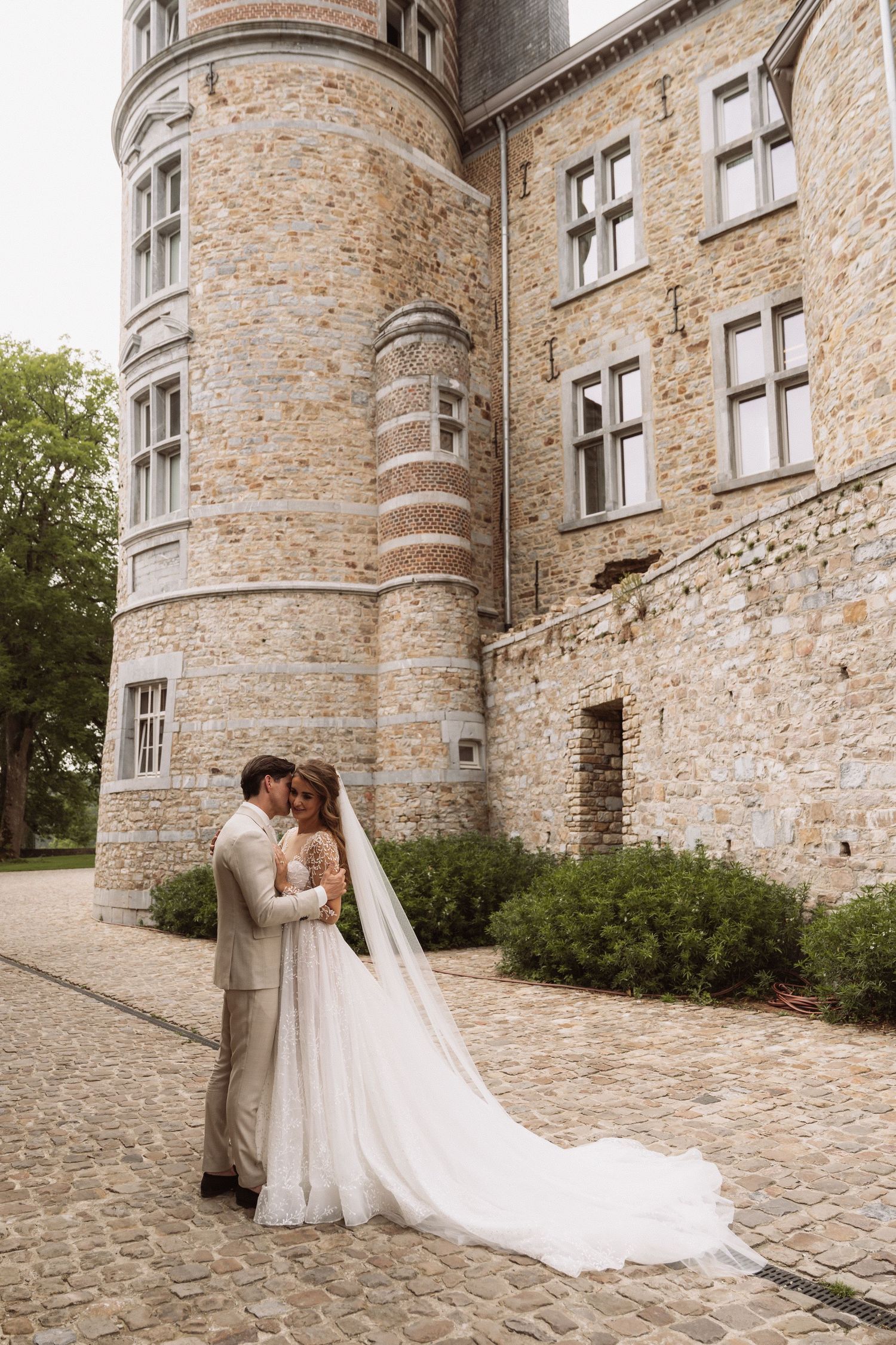Wedding couple poses against a historic stone castle wall with a flowing white dress train.