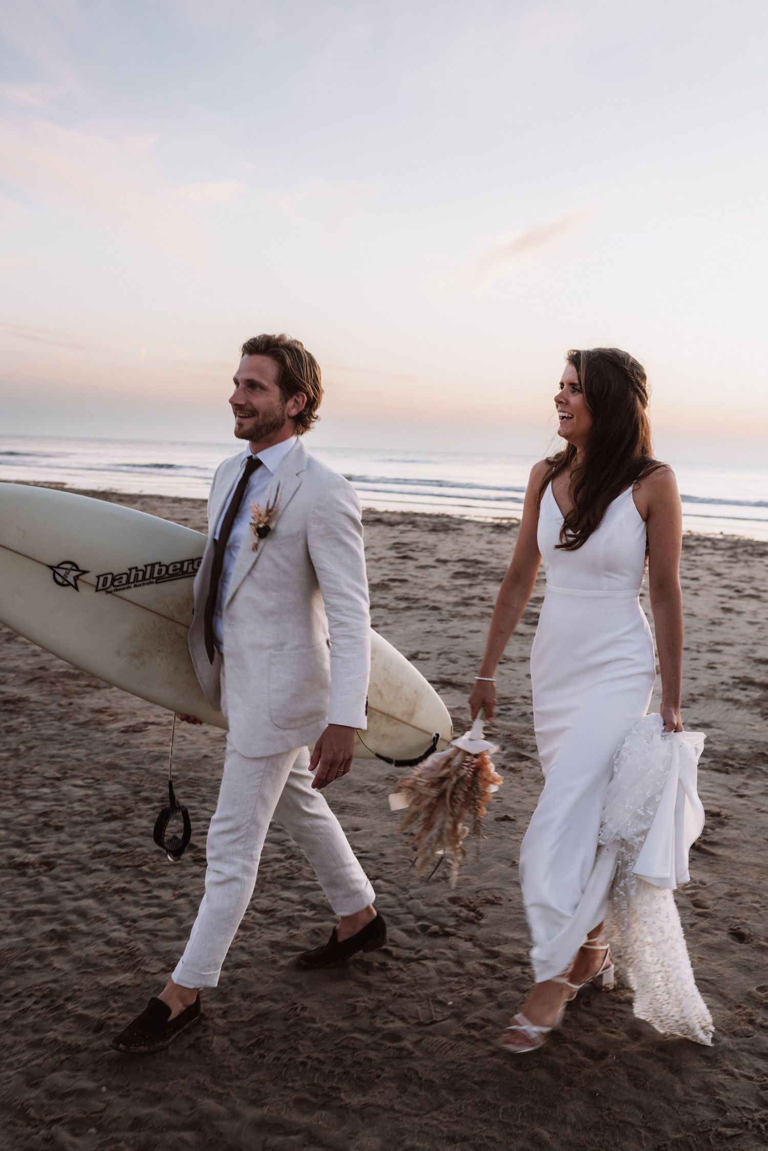 Newlyweds in white clothing walking on beach with surfboard during sunset.