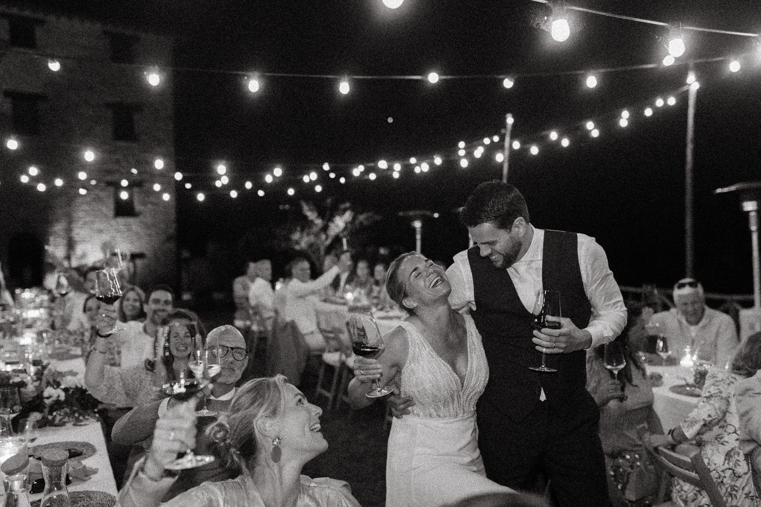 String lights illuminate an outdoor wedding reception with guests seated at long tables during an evening celebration.