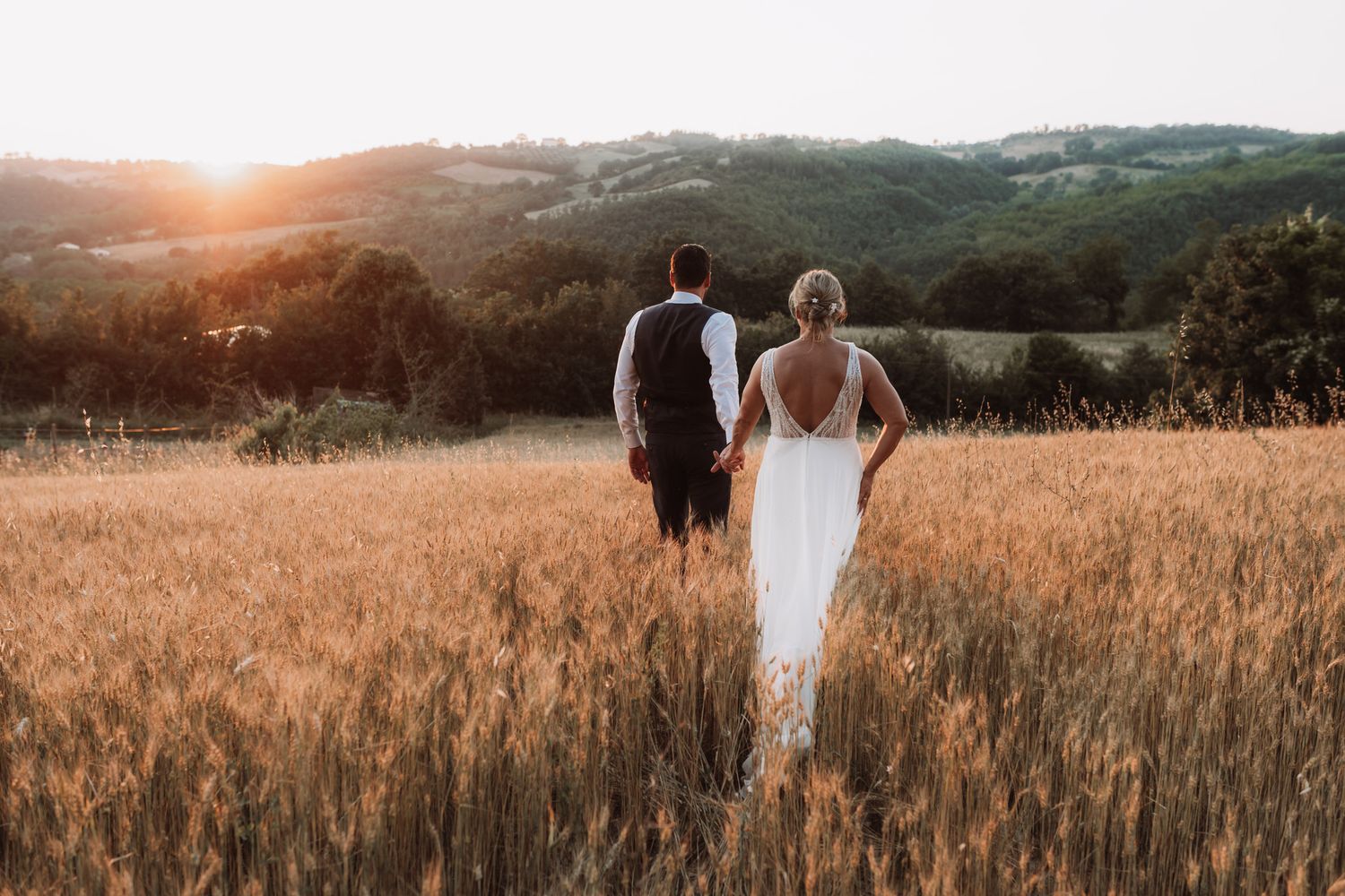 Couple in wedding attire walks through golden wheat field at sunset in countryside setting.