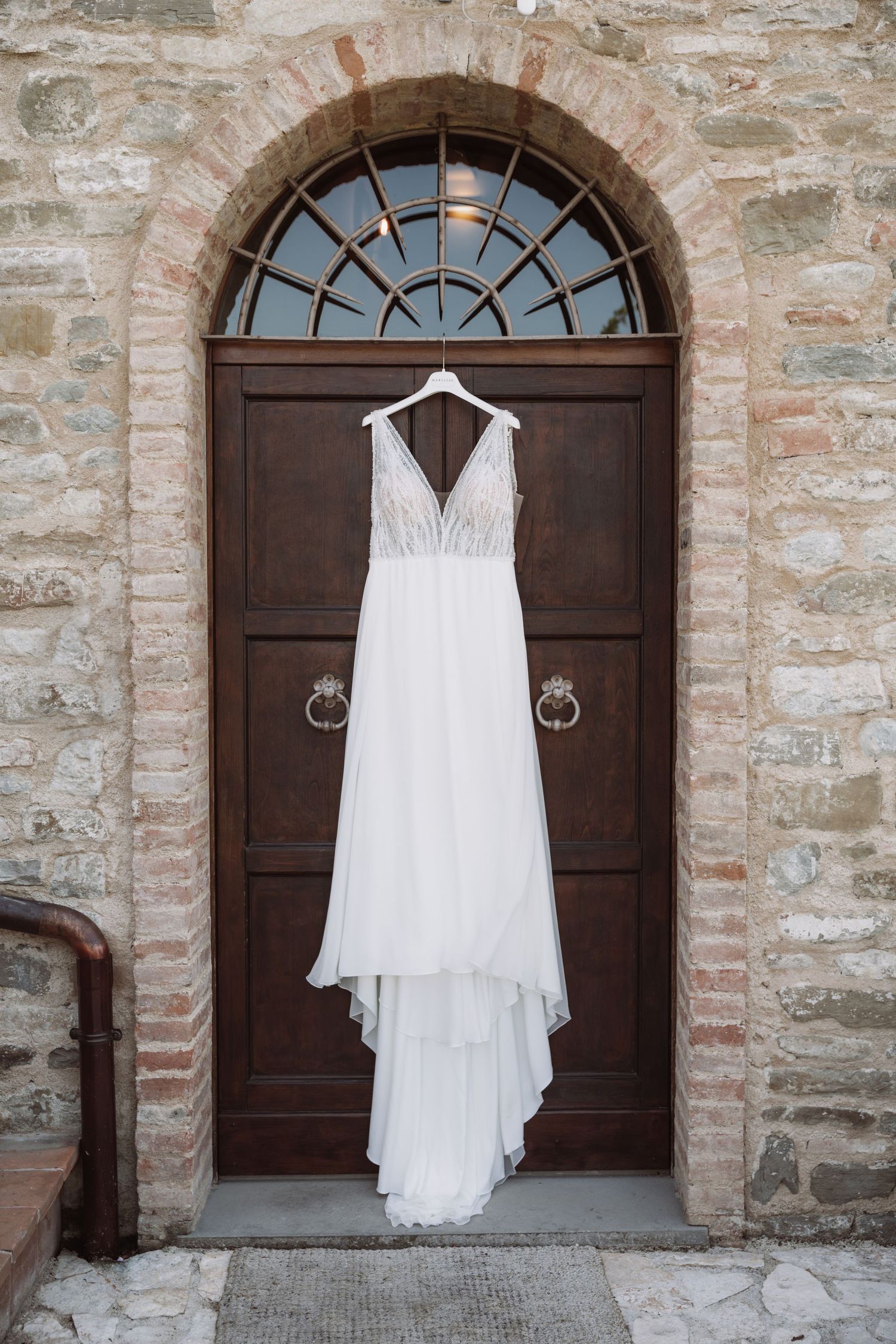 White wedding dress hanging against rustic stone wall with wooden arched door in elegant display.